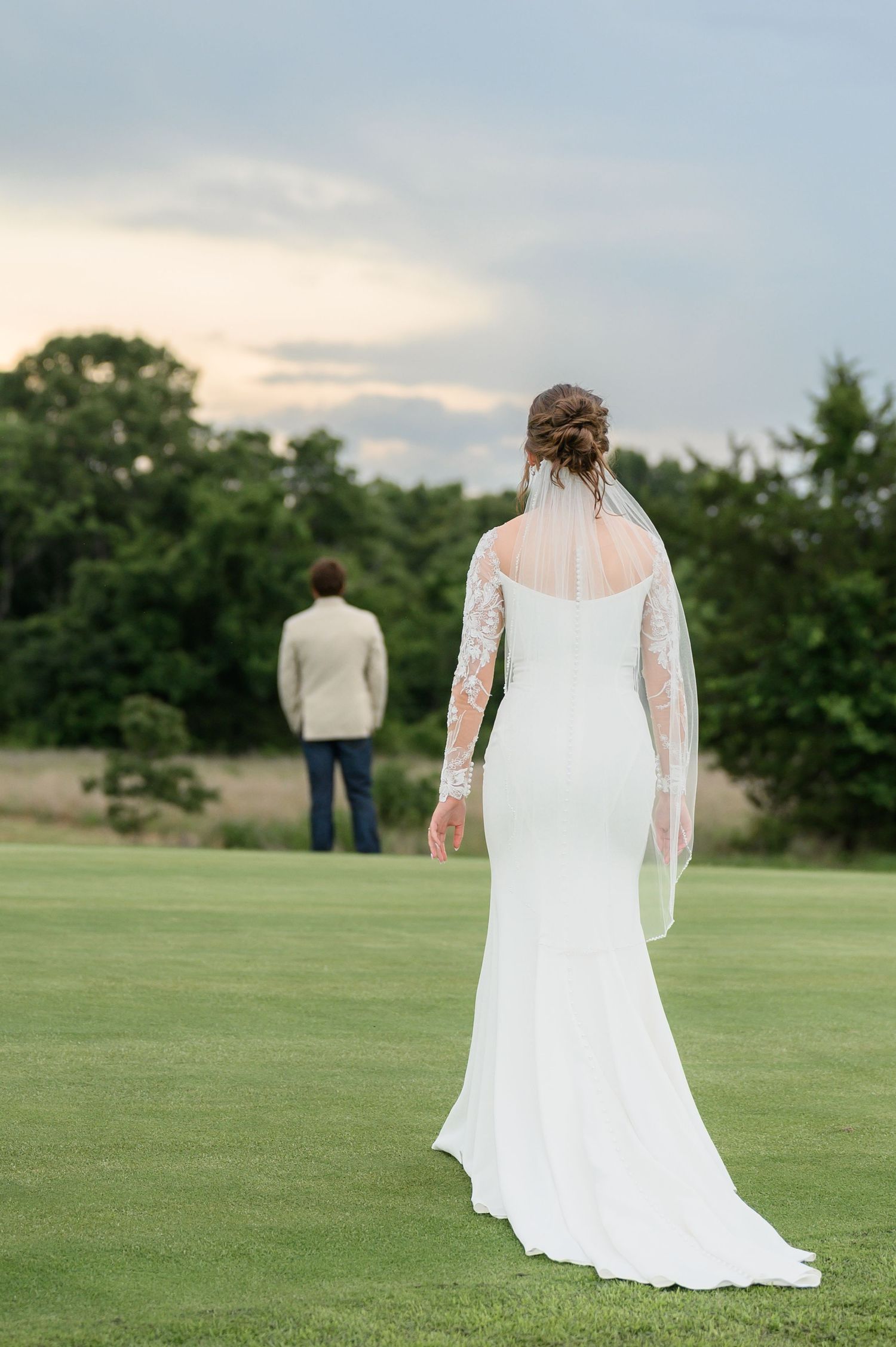 Bride in white lace gown with veil walks toward groom on green lawn at sunset.