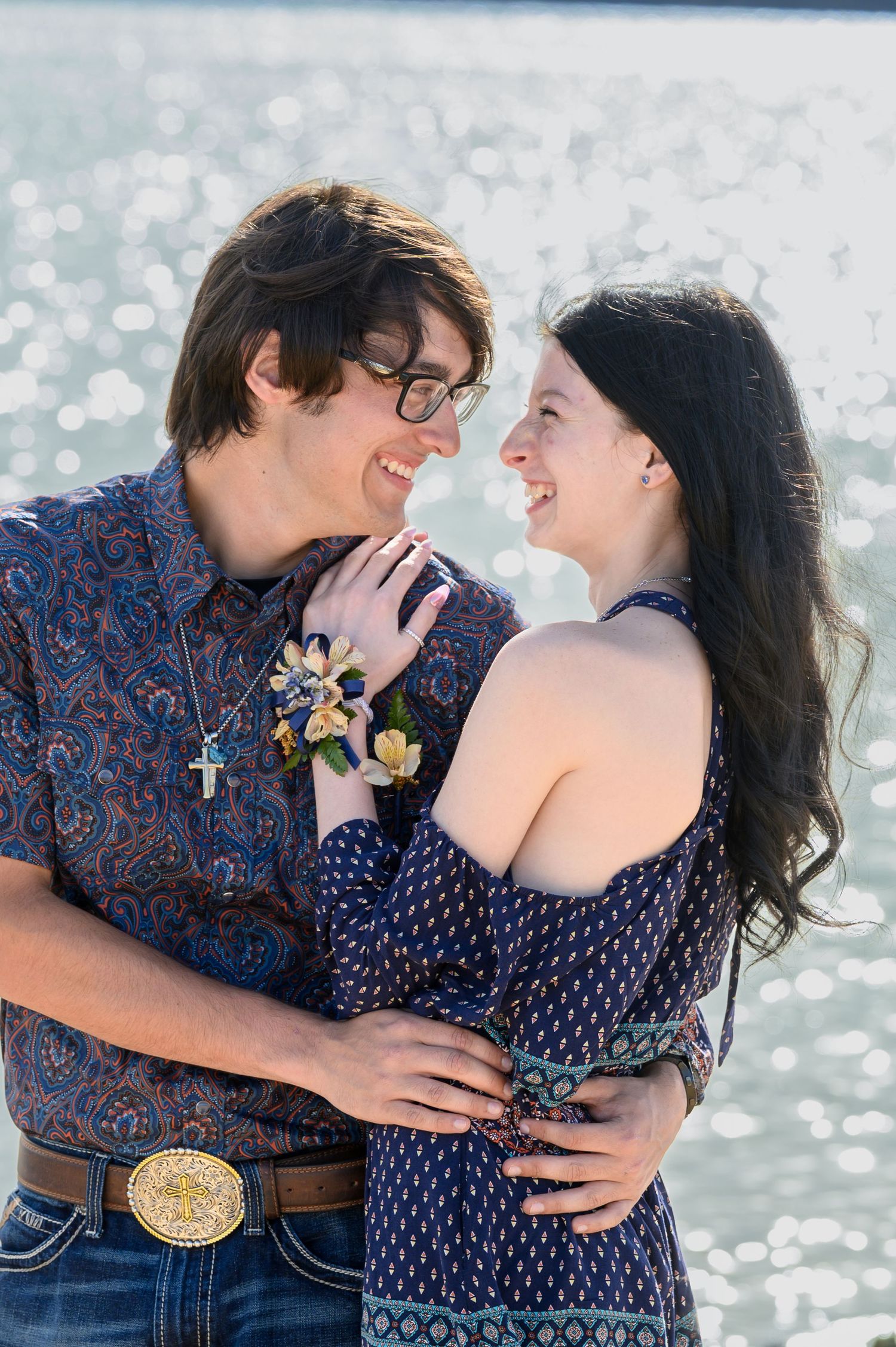 Smiling teenage prom couple holding each other near a shimmering lake.