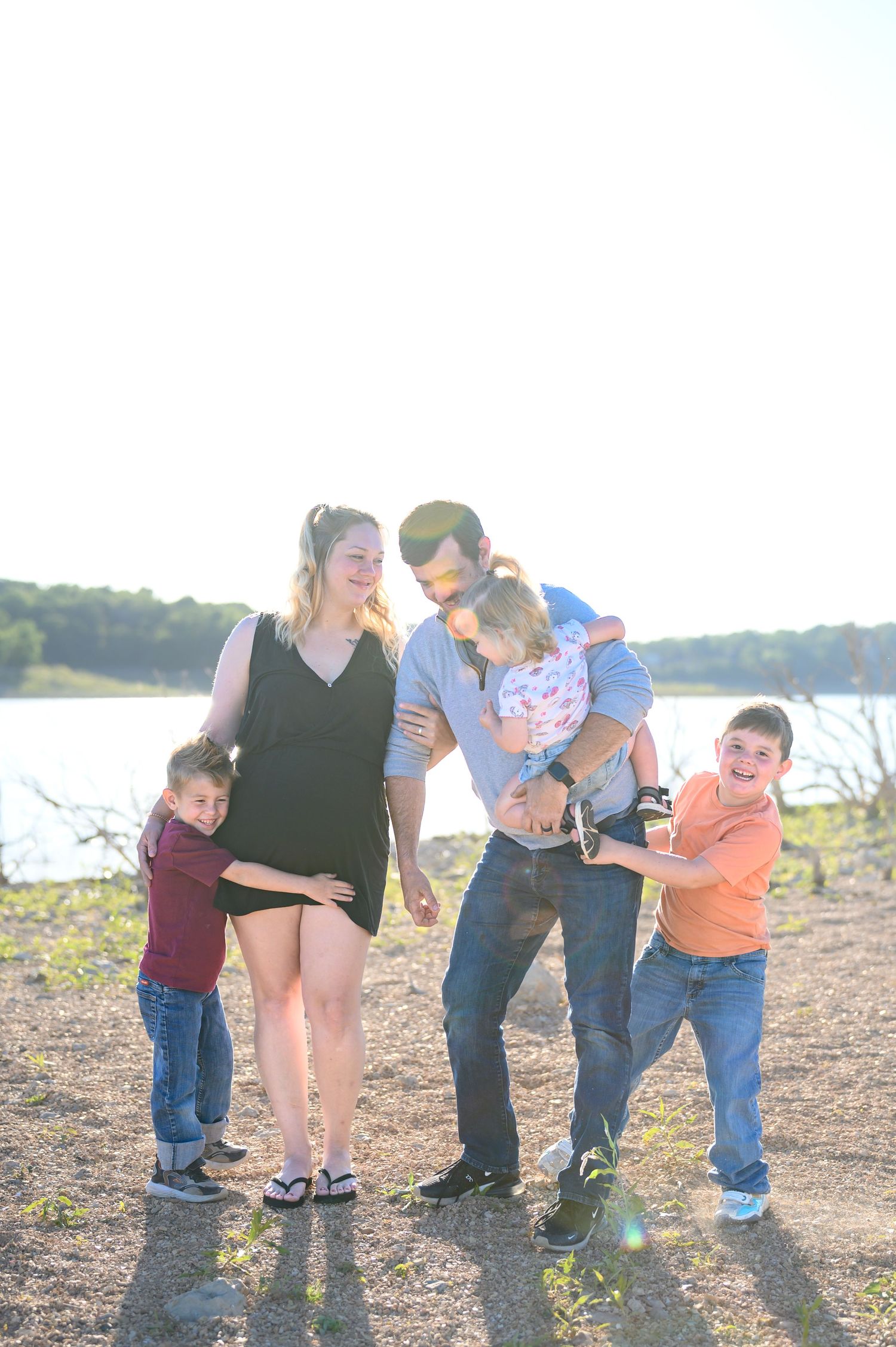 Playful family of five enjoying candid moment outdoors near lake with bright sunlight in background.