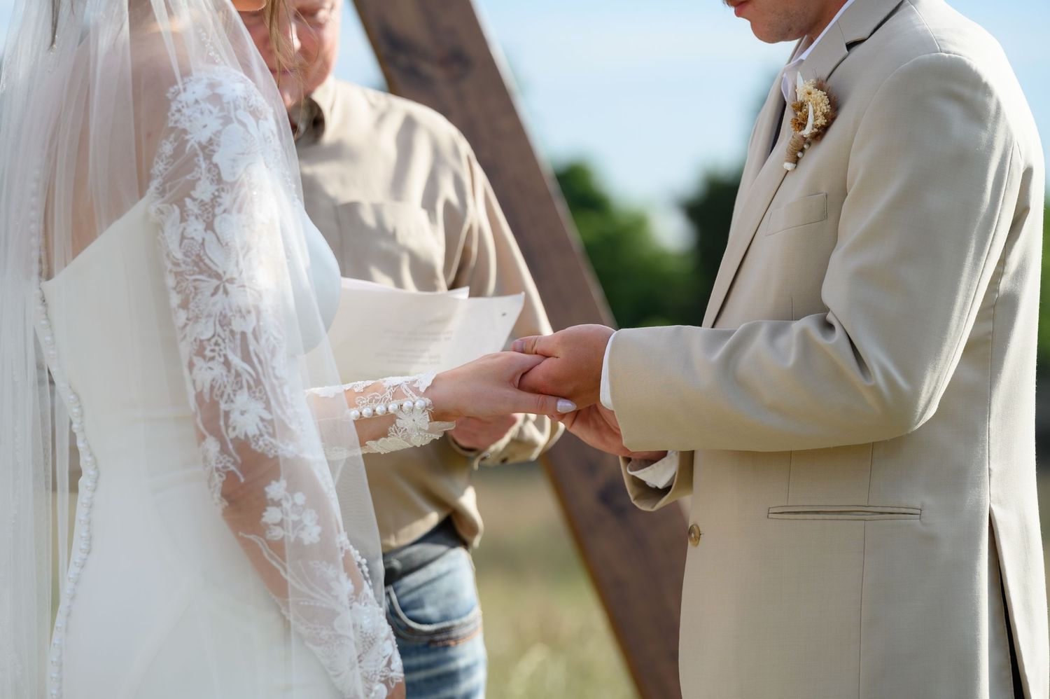 Groom in tan suit places wedding ring on brides lace-sleeved hand during outdoor ceremony.