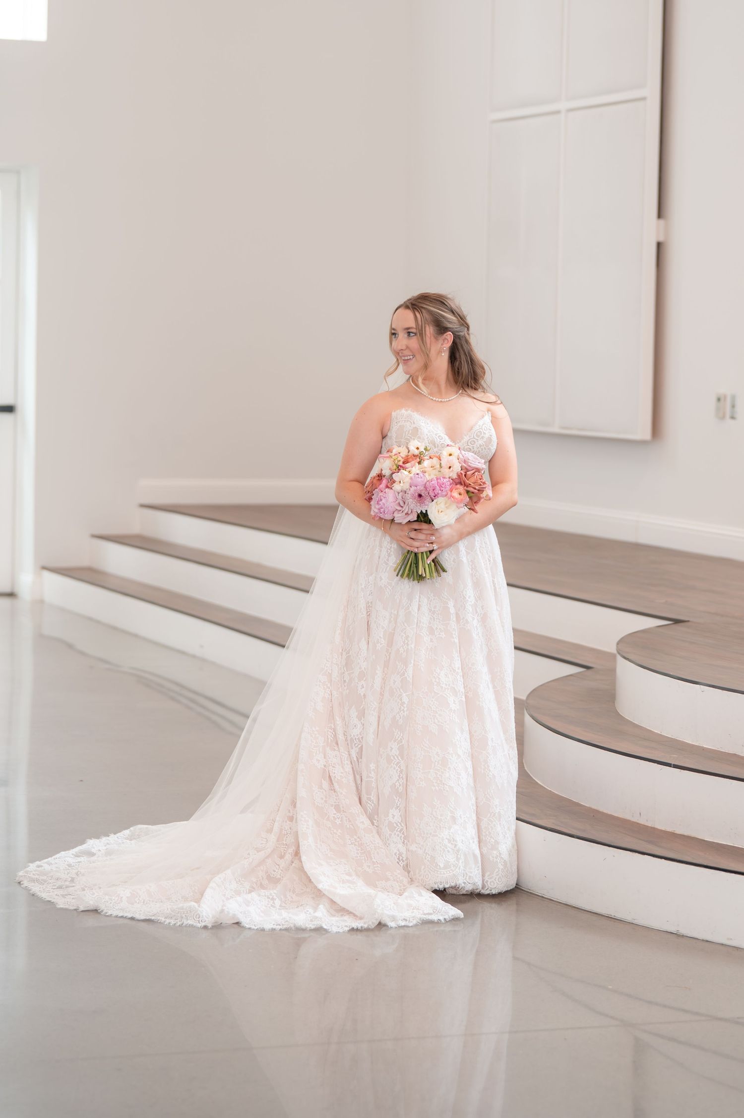 Elegant bride in strapless lace gown holds pink bouquet on marble staircase inside bright white venue.