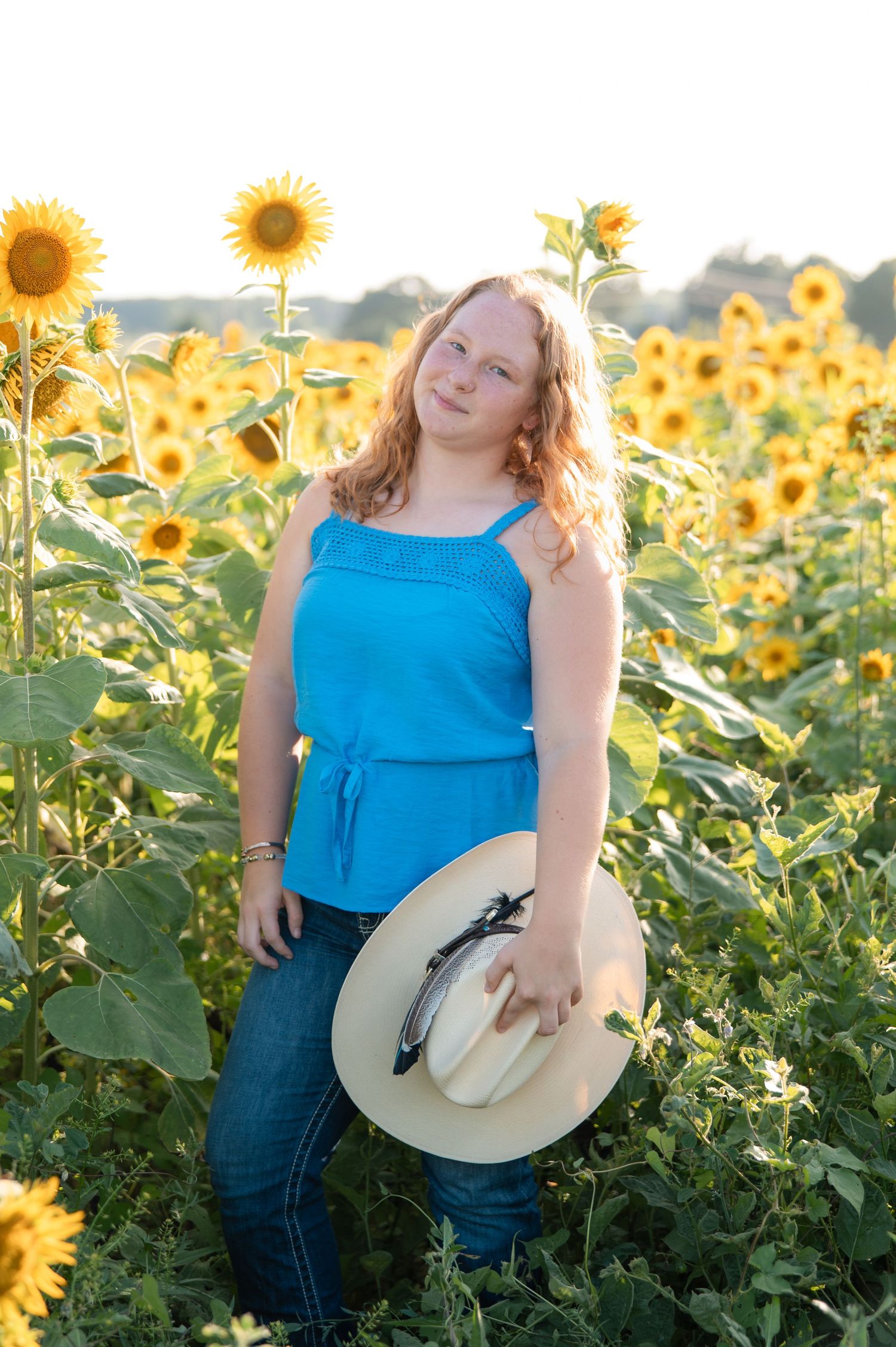Teen girl in blue tank top holding straw hat standing in a sunflower field at sunset.