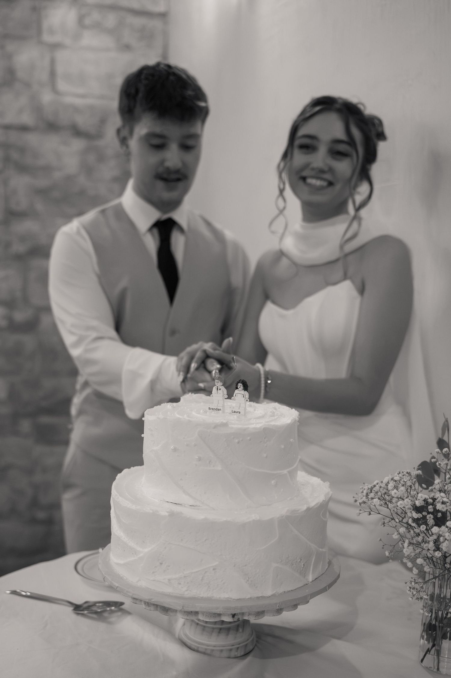 Bride and groom cutting their two-tier white wedding cake topped with figurines in a black and white photo.