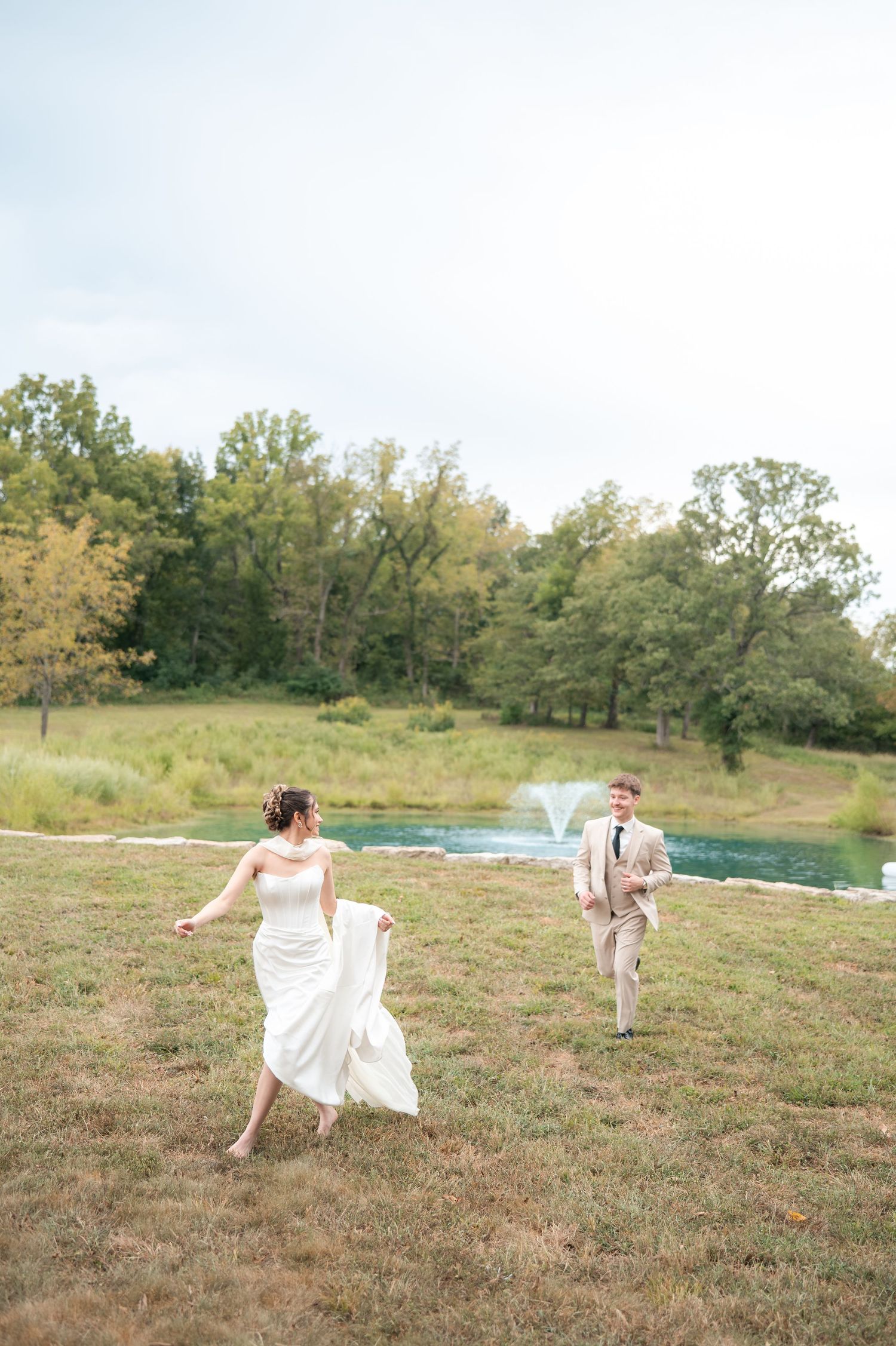 Bride in white gown and groom in tan suit running joyfully across green lawn with pond and fountain backdrop.