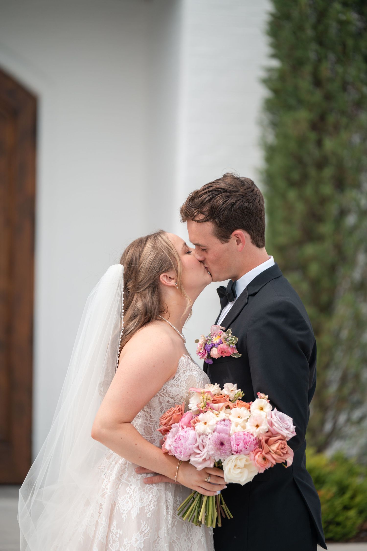 Bride and groom share romantic kiss holding colorful pink and coral bouquet outside white wedding venue.