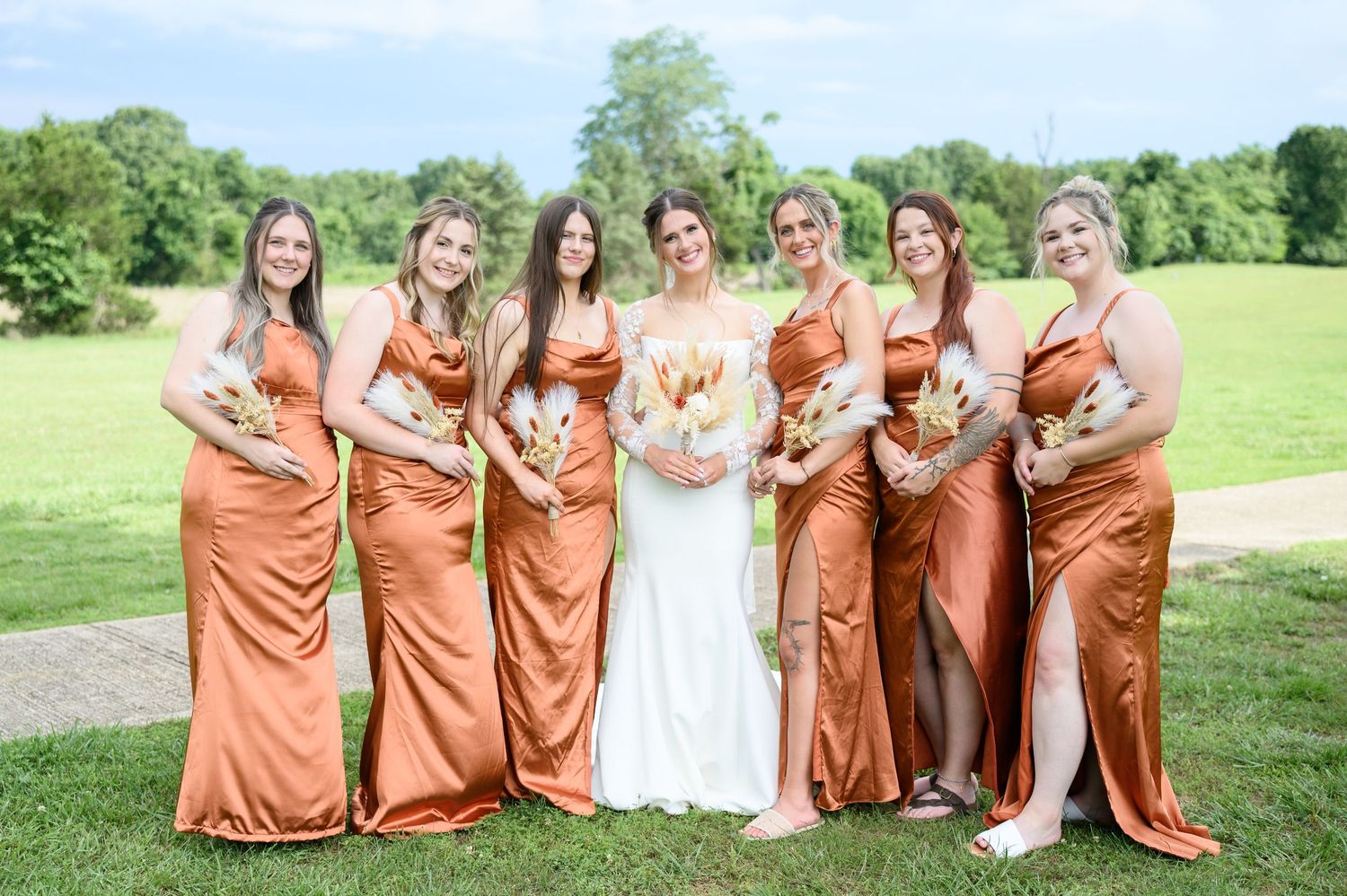 Bride in white gown poses with six bridesmaids wearing rust orange satin dresses holding dried floral bouquets outdoors.