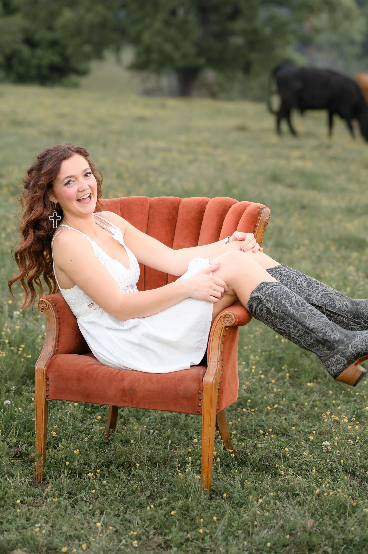 Young woman in white dress and tall boots laughing while seated in a velvet chair outdoors.