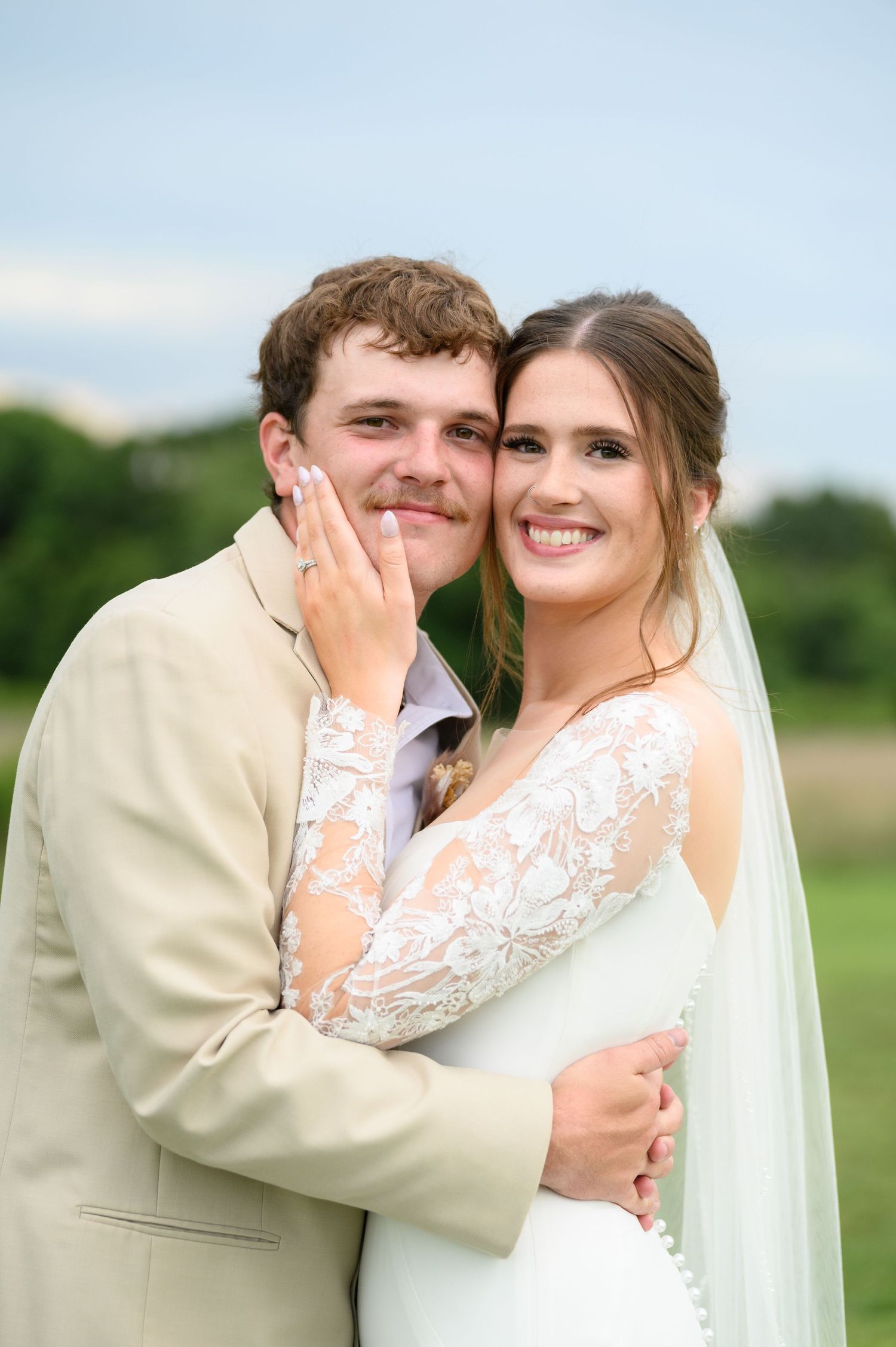 Smiling bride and groom embracing closely outdoors, bride wearing lace long-sleeve wedding gown with veil.