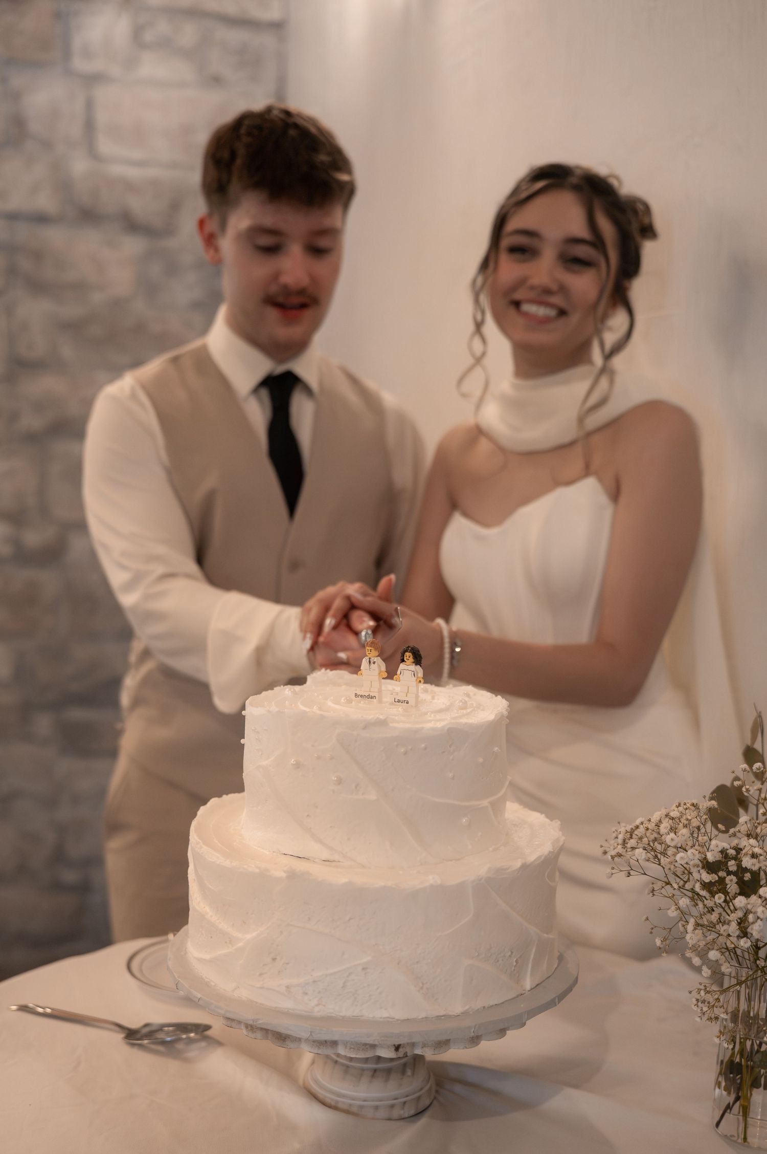 Bride and groom smiling while cutting a two-tier white wedding cake topped with miniature figurines.