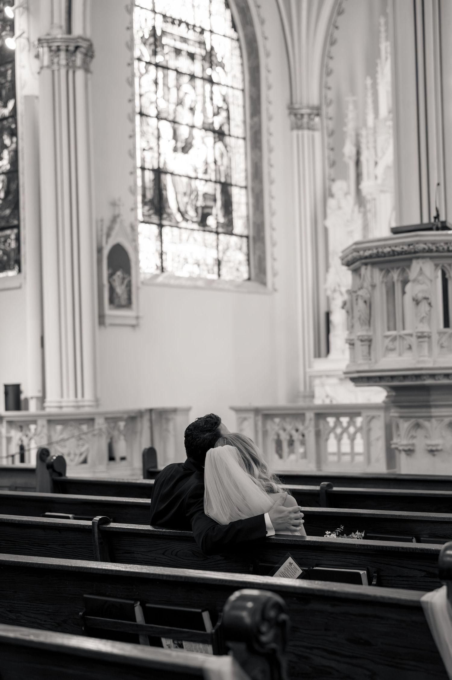 Bride and groom sit intimately together on a church pew beneath stunning stained glass windows in black and white.