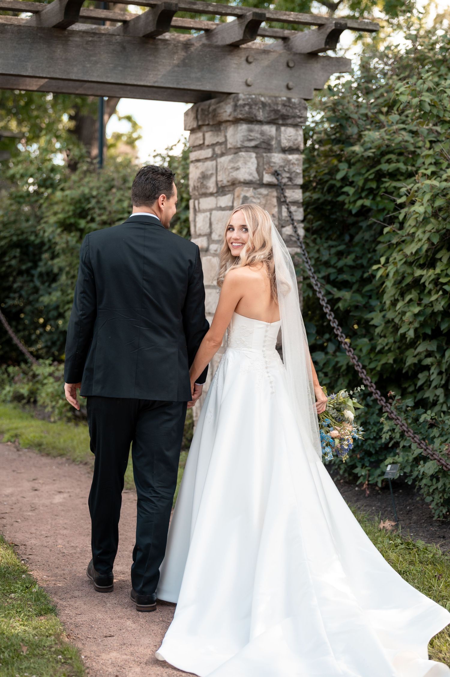 Bride and groom walk hand in hand through lush garden pergola, bride in elegant white gown.