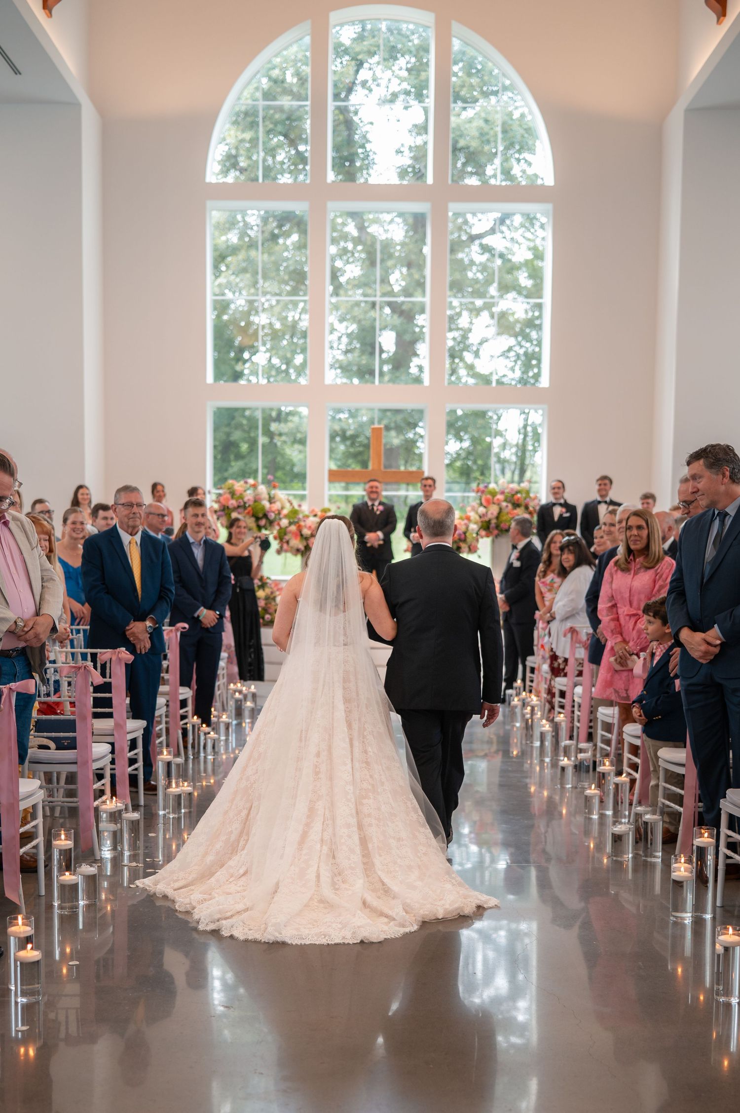 Bride walks down aisle in lace gown with long veil in sunlit chapel filled with wedding guests.