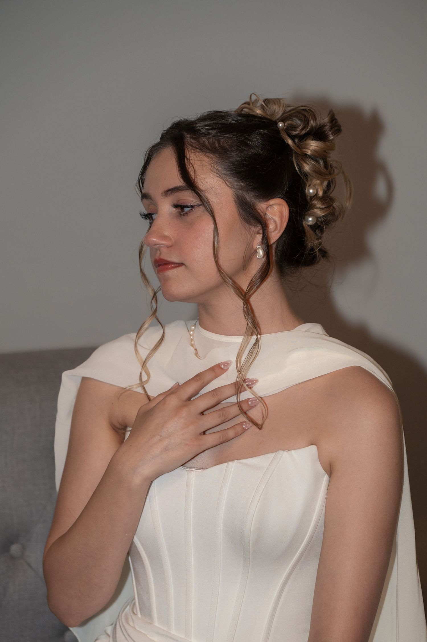 Young woman in white gown with elegant updo hairstyle, pearl earrings, and hand placed gracefully on her chest.