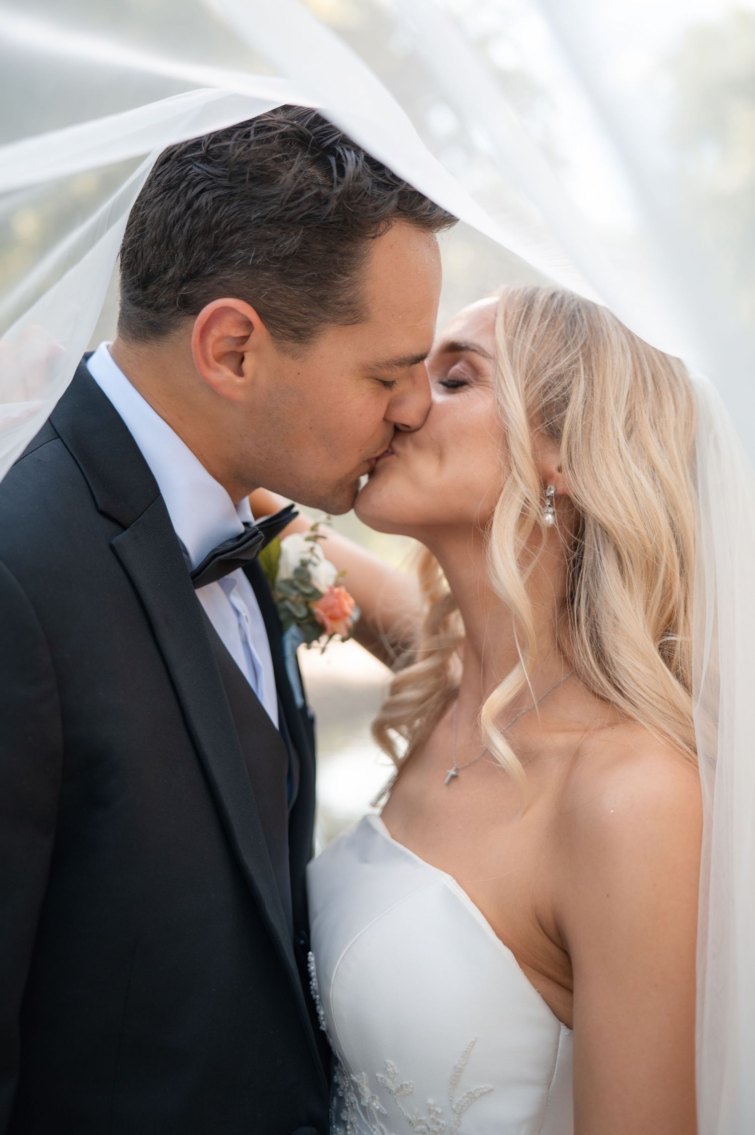Bride and groom share romantic kiss under flowing white veil in soft natural light.