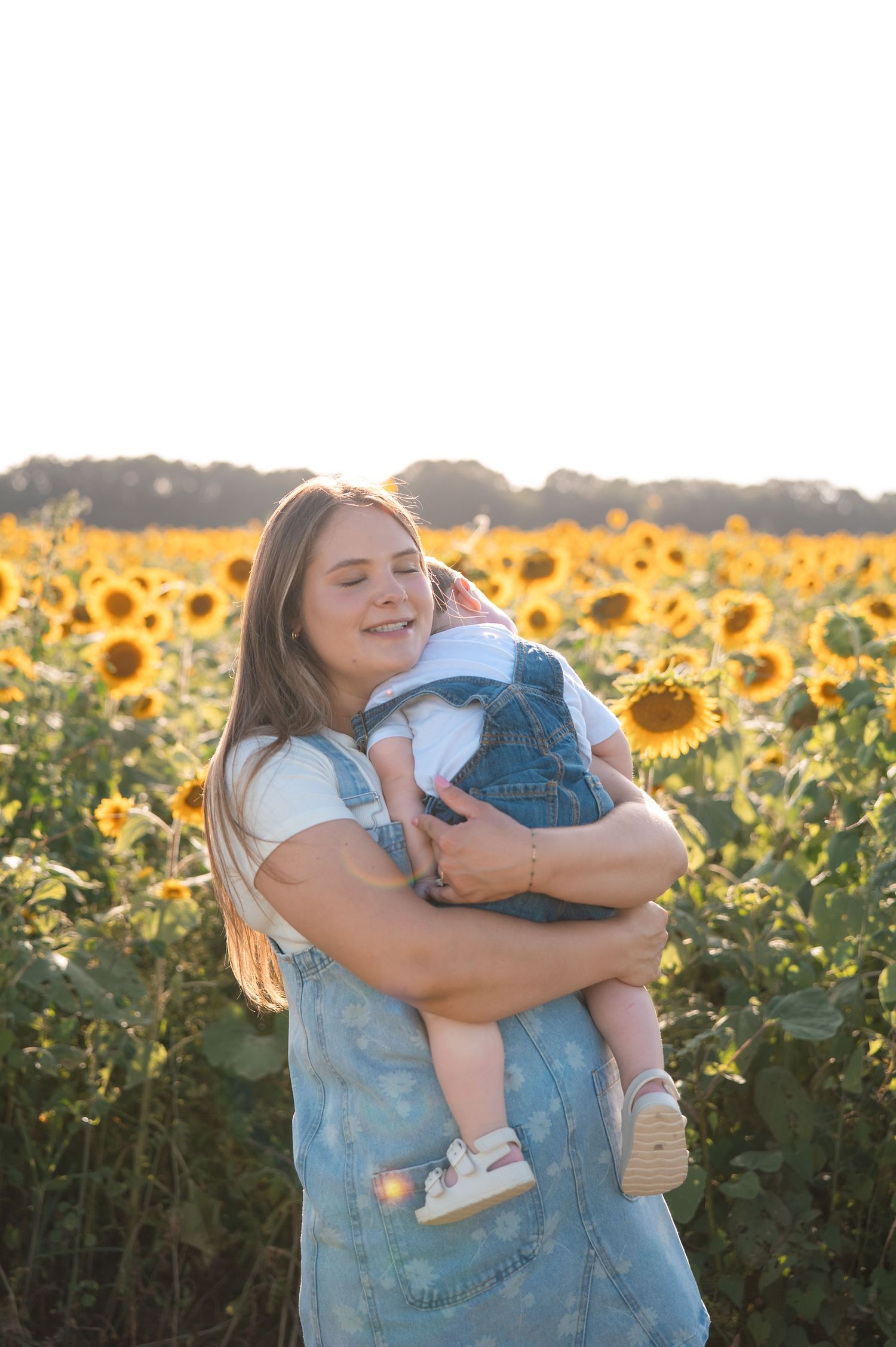Smiling mother in denim dress holding baby in sunflower field bathed in warm sunset light.