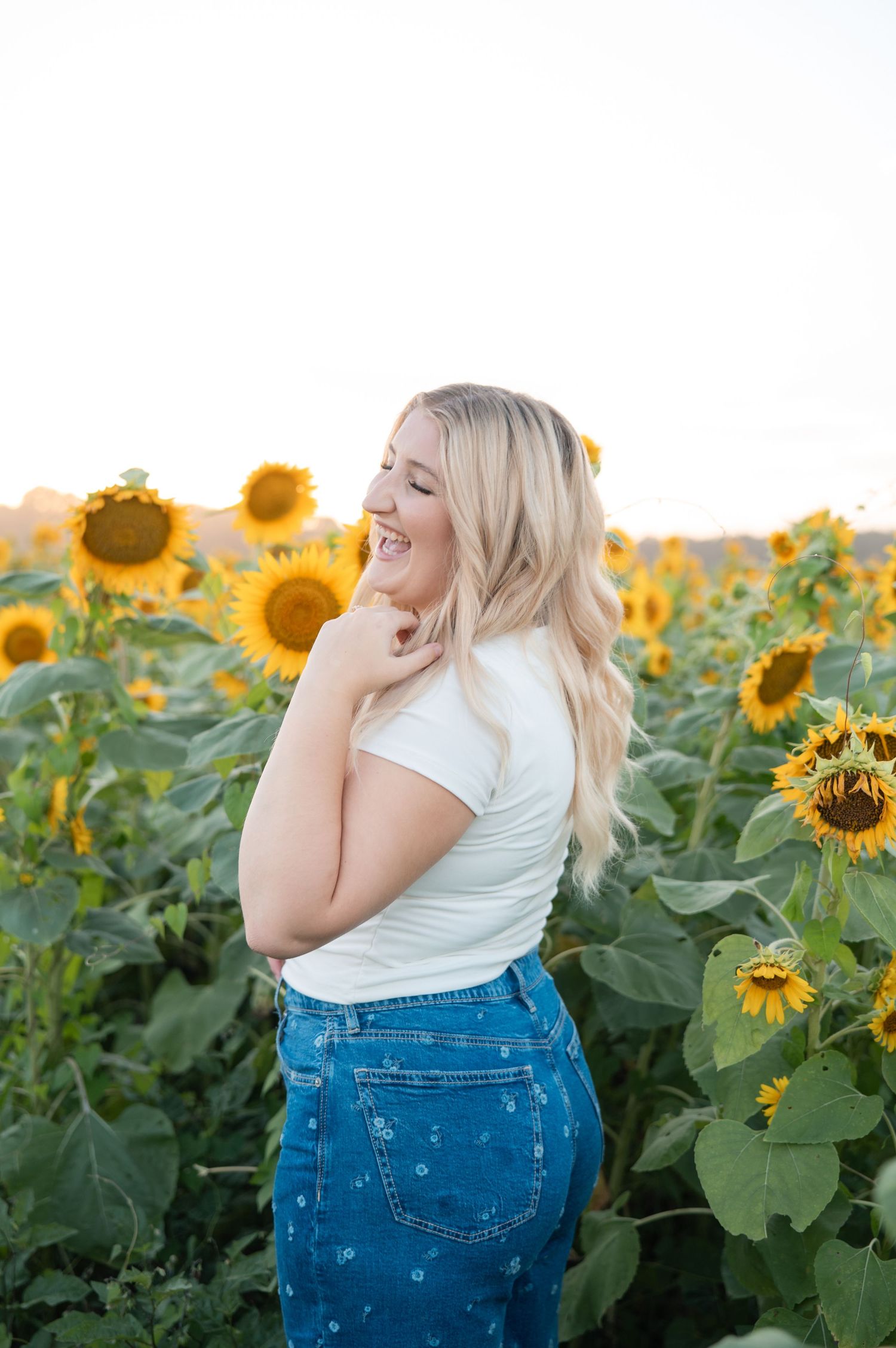Blonde woman laughing in sunflower field at sunset, wearing white tee and floral denim skirt.
