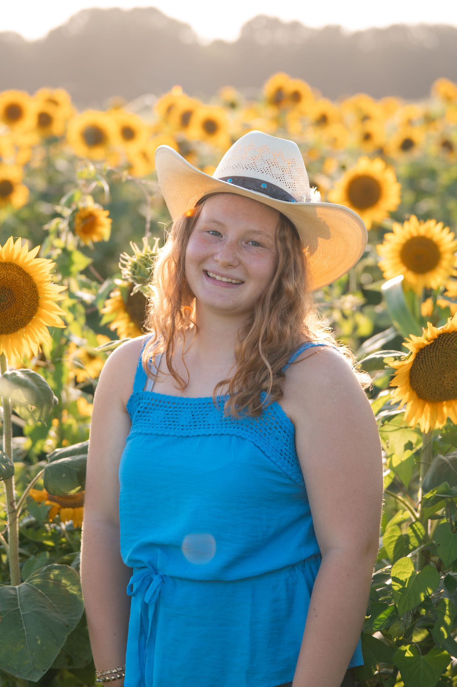 Smiling senior wearing cowboy hat and blue dress standing in vibrant sunflower field at sunset.