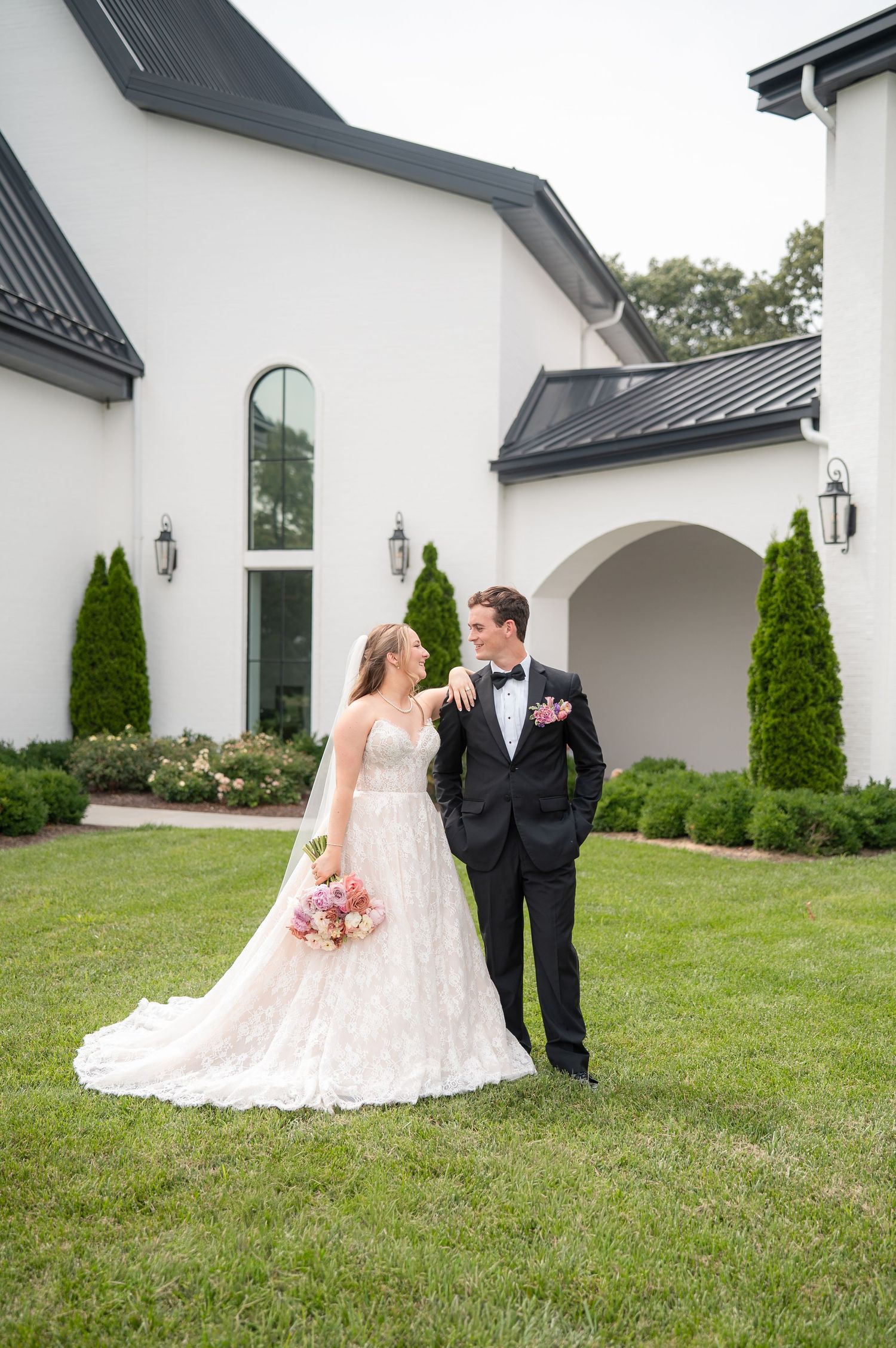 Bride and groom share a loving glance on lush lawn outside white modern wedding estate.