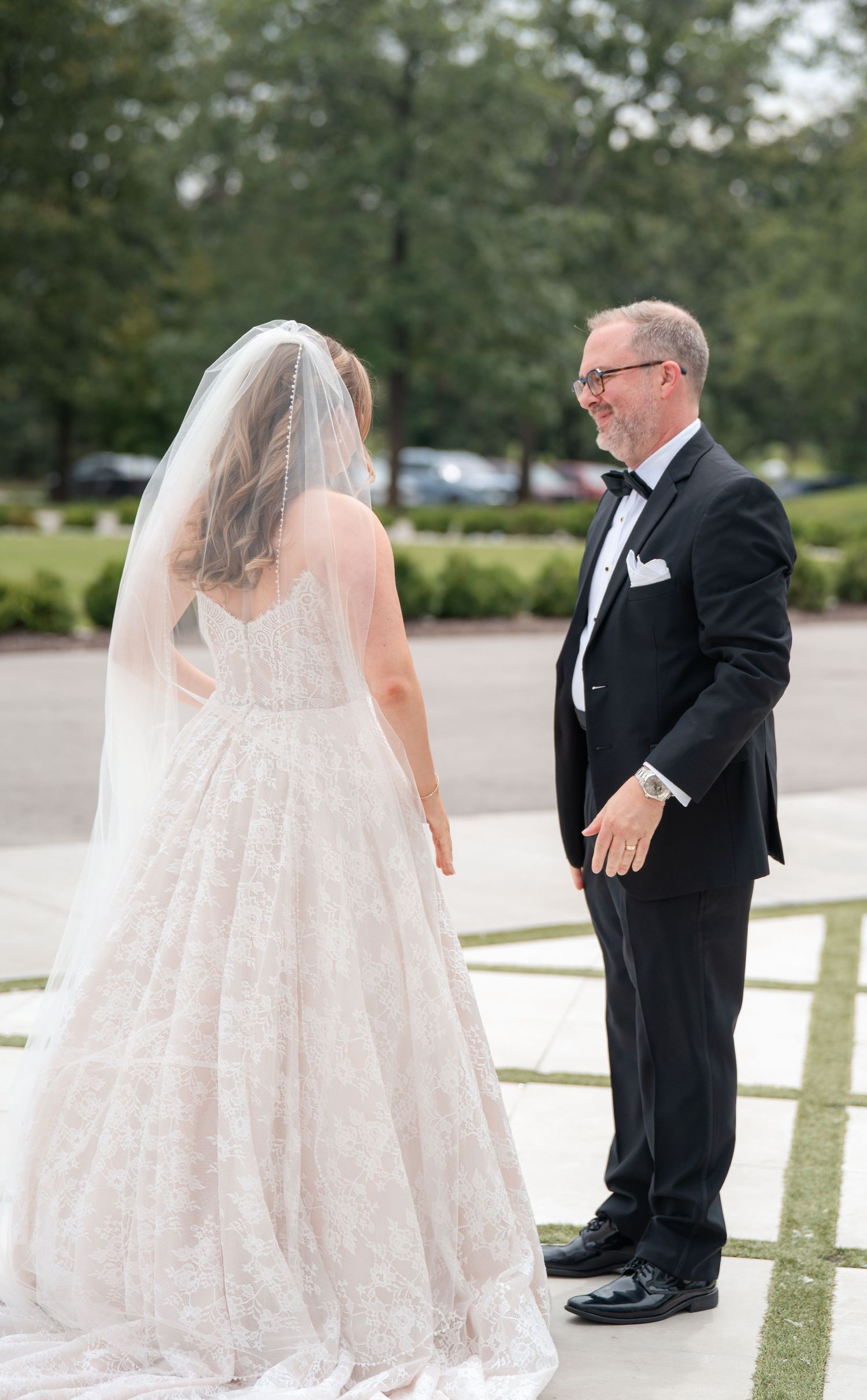 Father smiling at bride in lace gown with veil during first look moment.