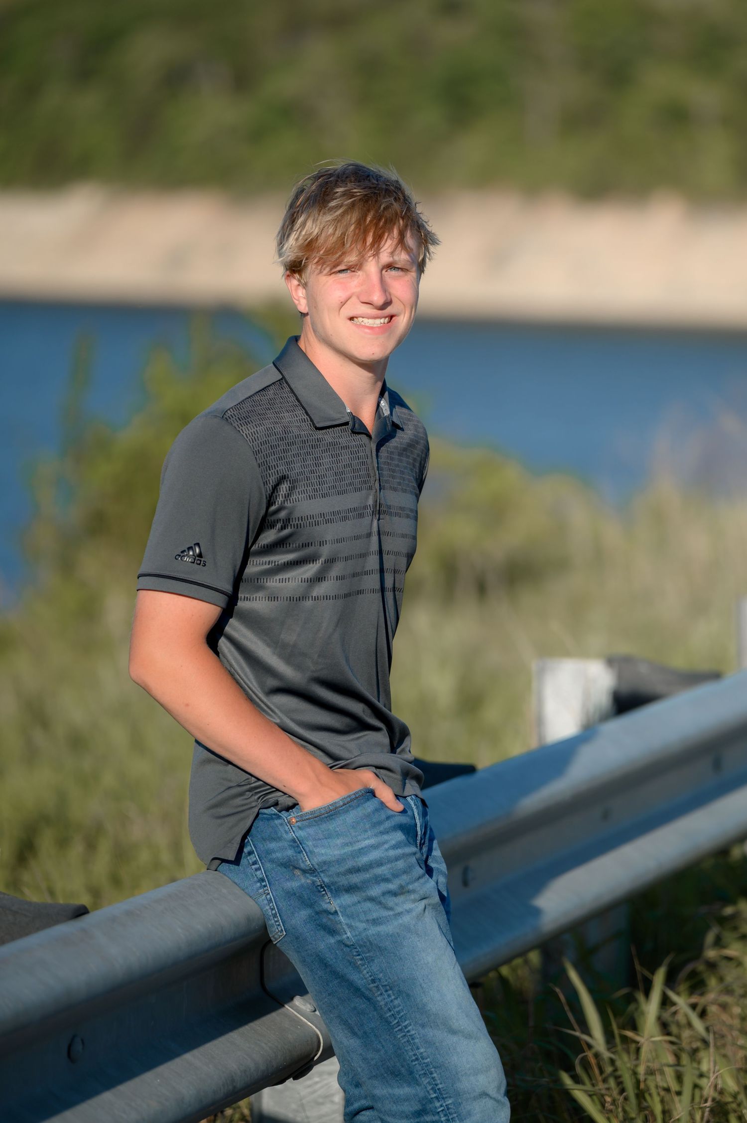 Smiling blonde teen boy in gray Adidas polo leaning on a guardrail near a river at sunset.
