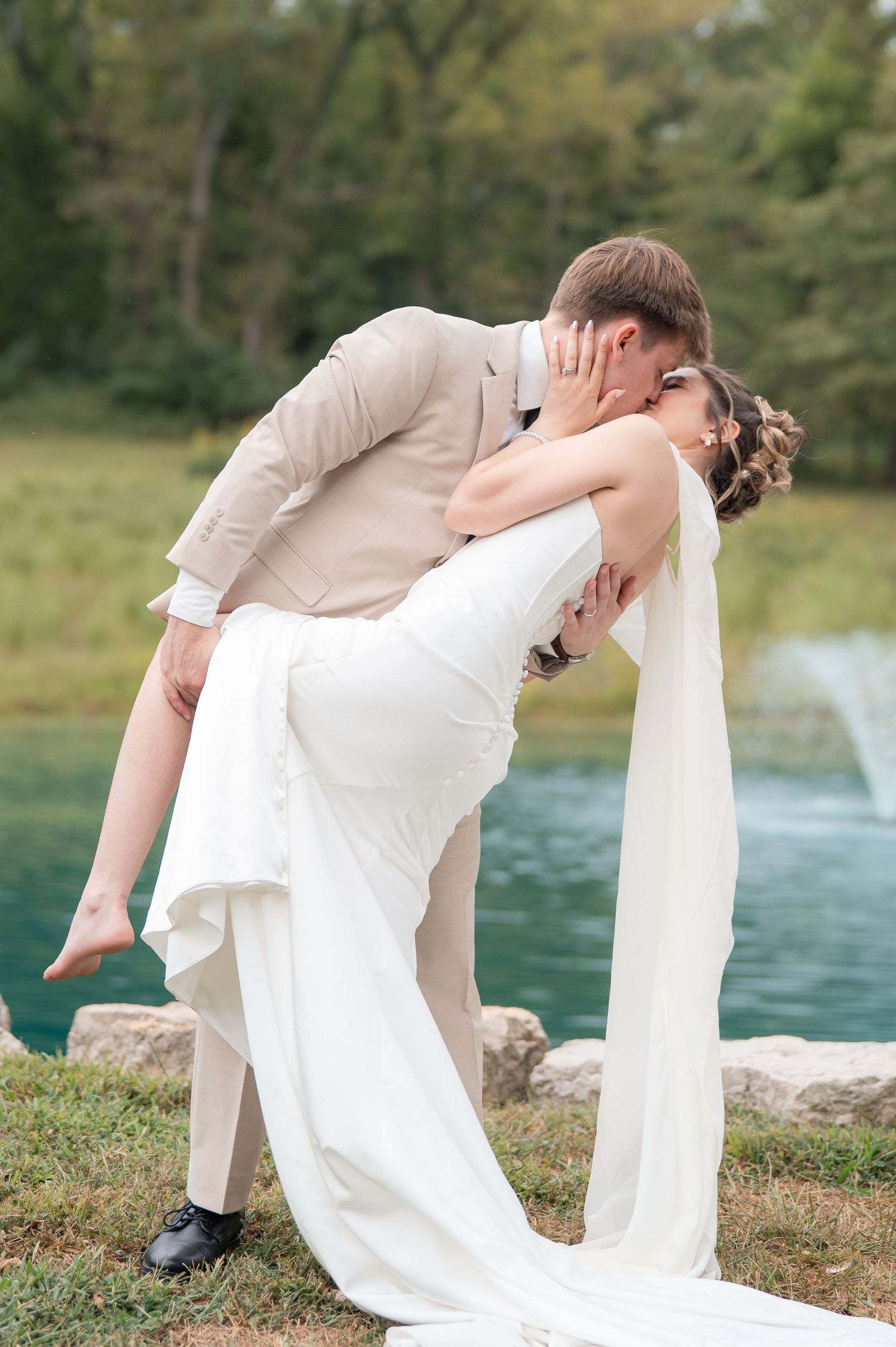 Groom dips bride in elegant white gown for a romantic kiss outdoors near a scenic pond.