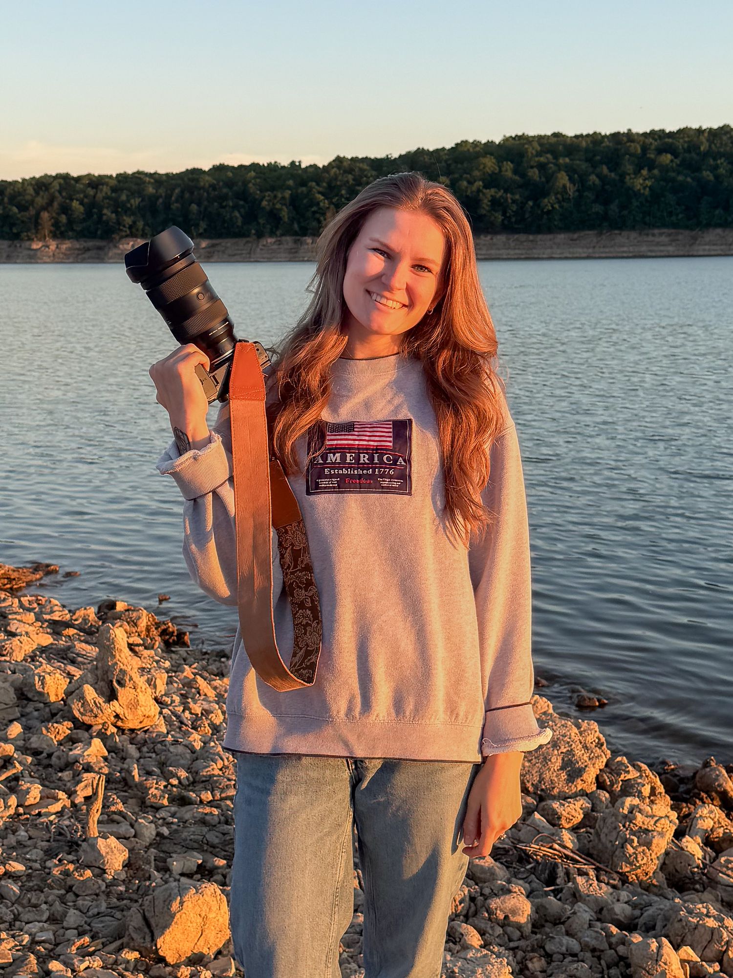 Young woman smiling outdoors holding a camera with a leather strap near a lake at golden hour.