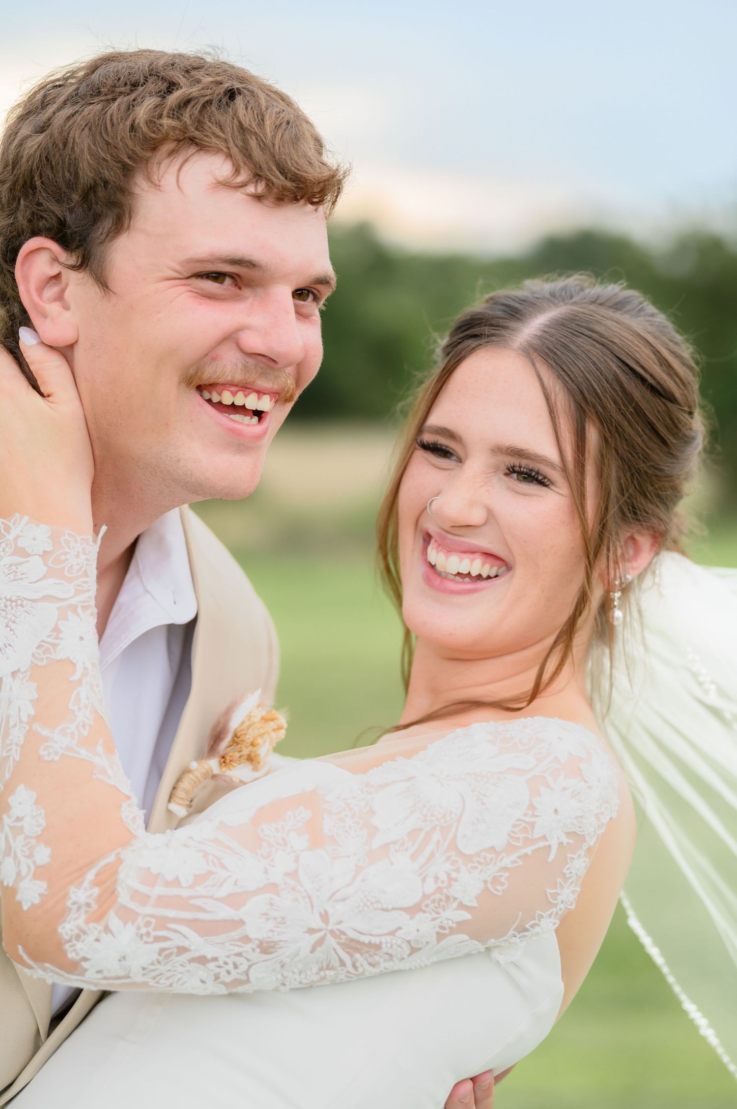 Bride and groom laugh together in a close embrace on a sunny outdoor wedding day.