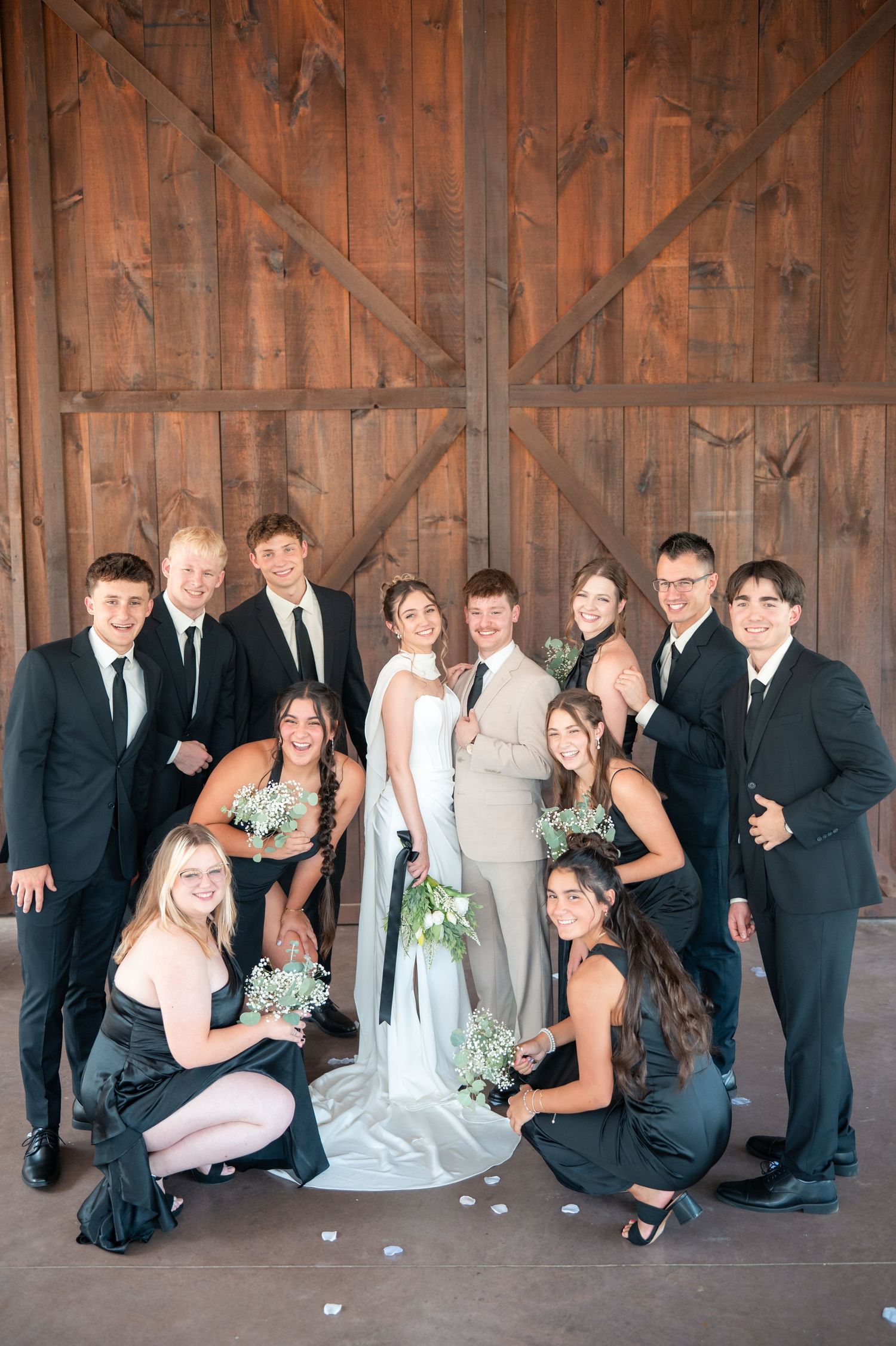 Wedding party poses in front of rustic barn doors, bride and groom centered among groomsmen in black suits and bridesmaids in dark gowns.