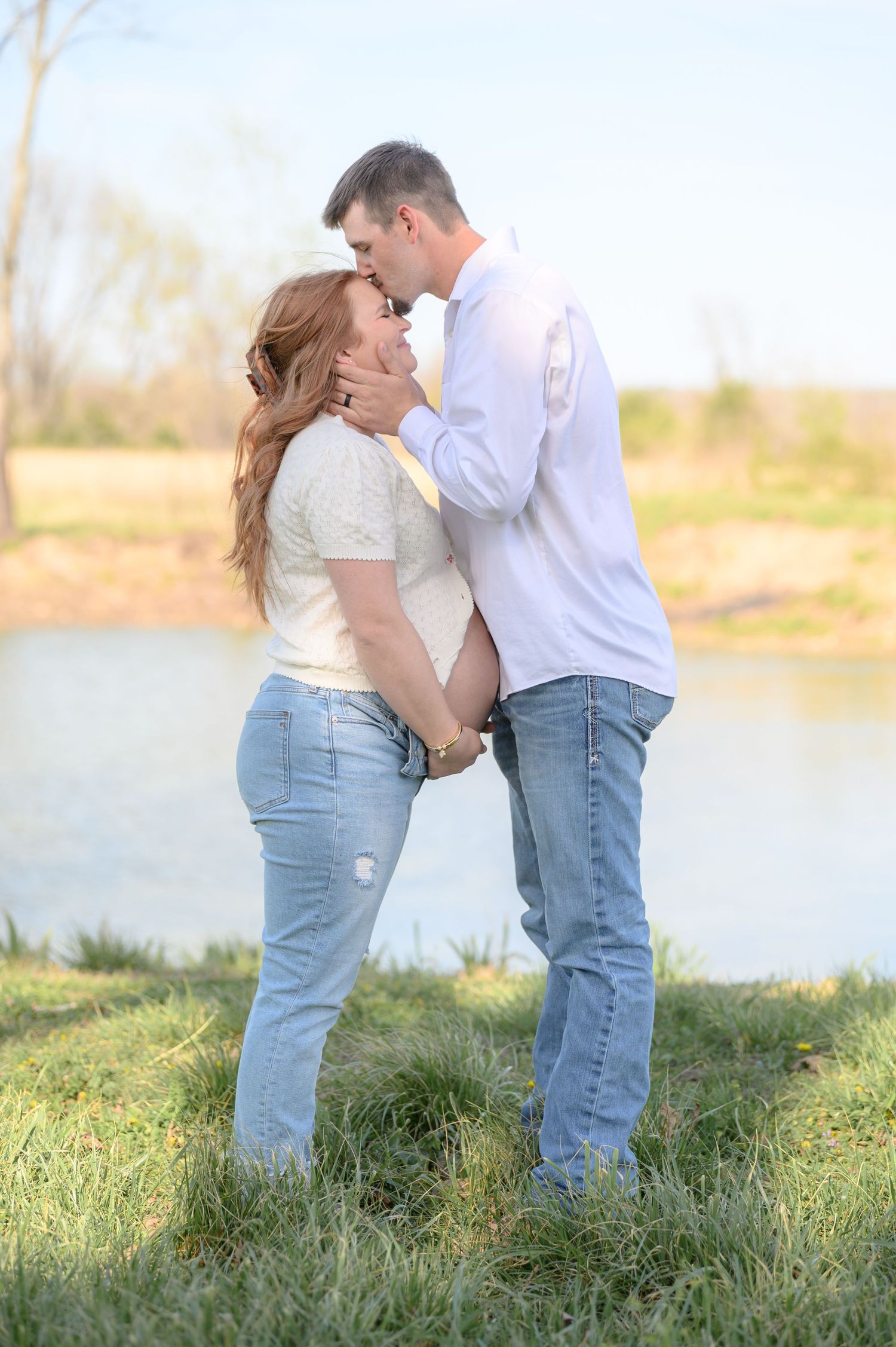 Expecting couple sharing a kiss near a serene pond at sunset.