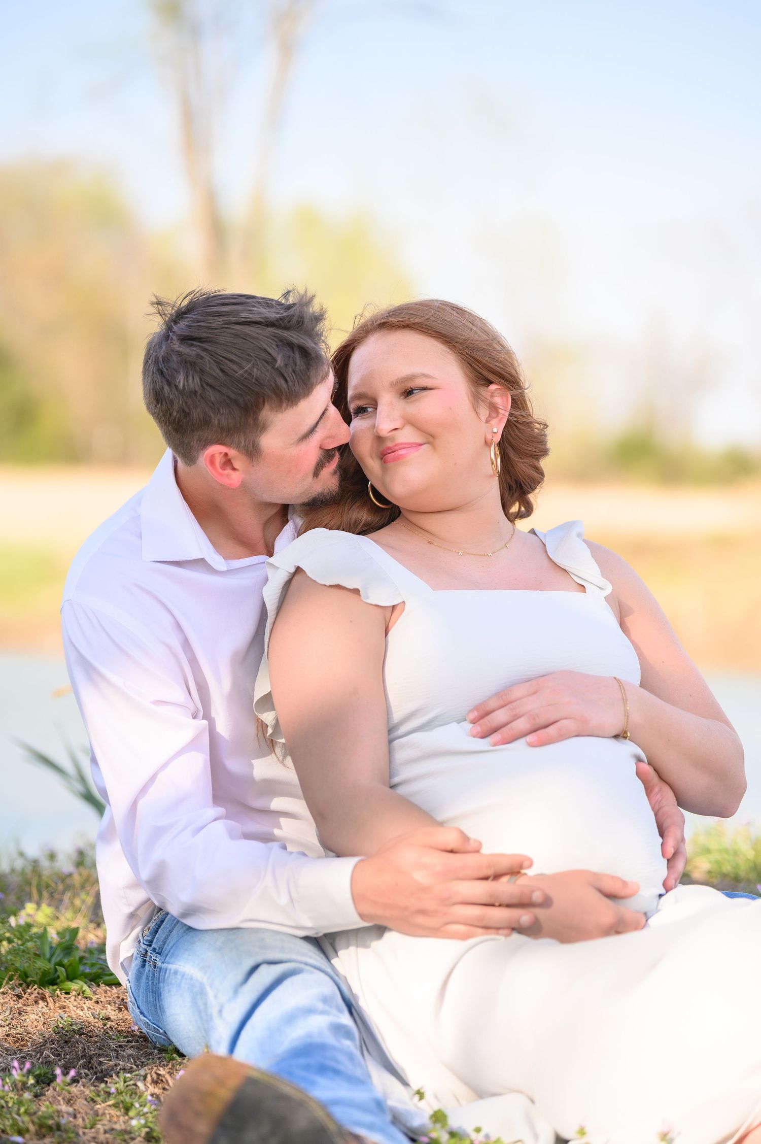 Couple sitting outdoors by water, man kissing pregnant woman's head while holding her baby bump in golden light.