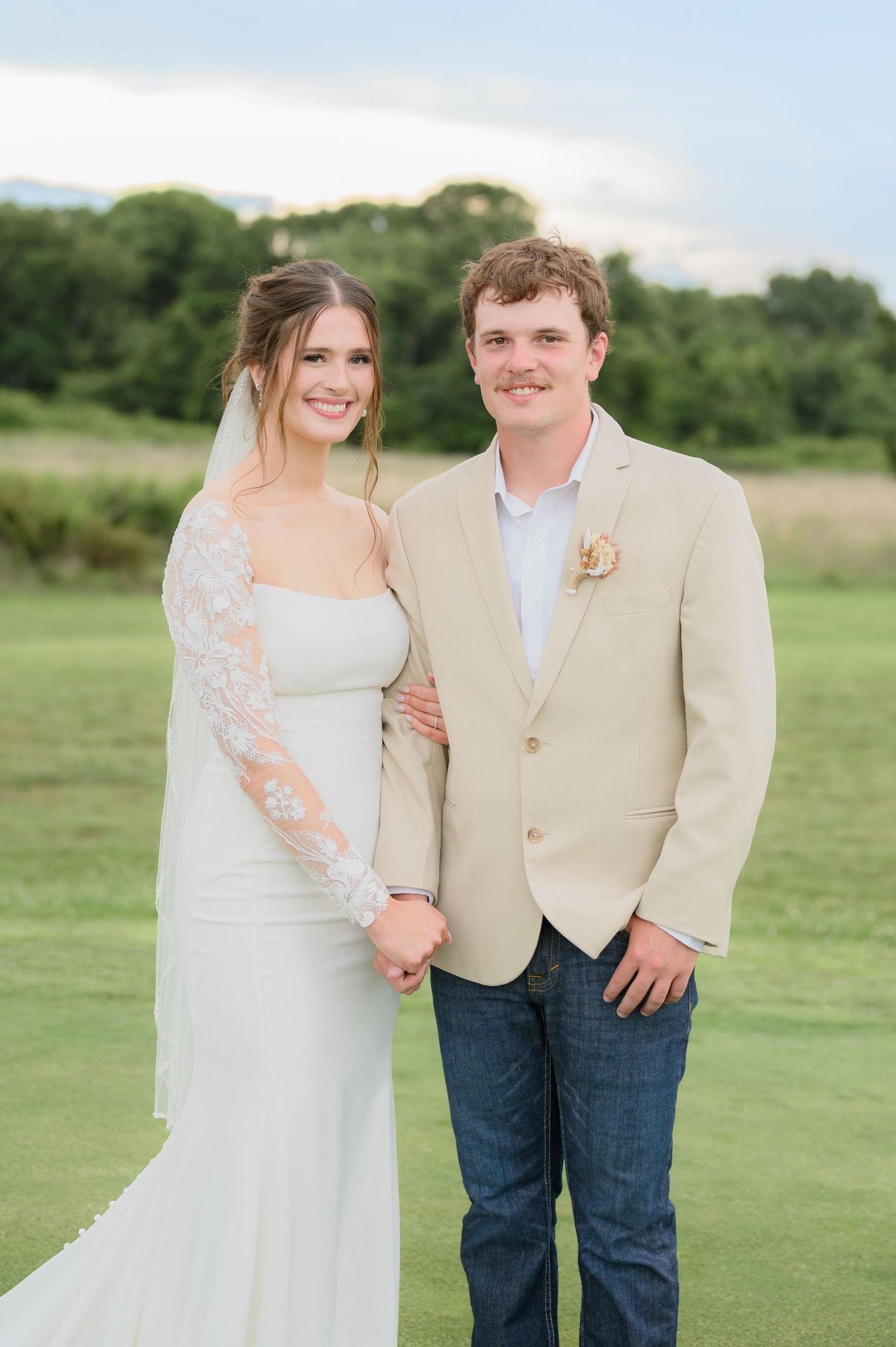 Bride and groom smile together on green lawn, bride in lace gown, groom in tan blazer.