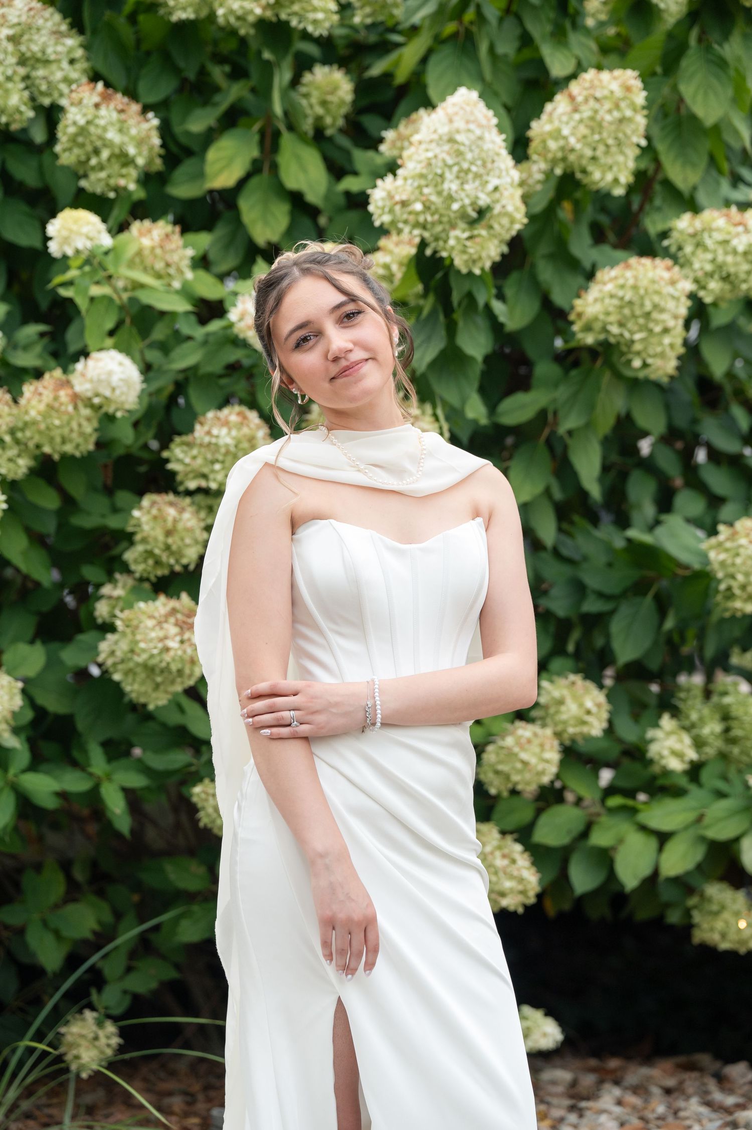 Bride in white strapless gown with draped shoulder detail poses before lush hydrangea bushes.