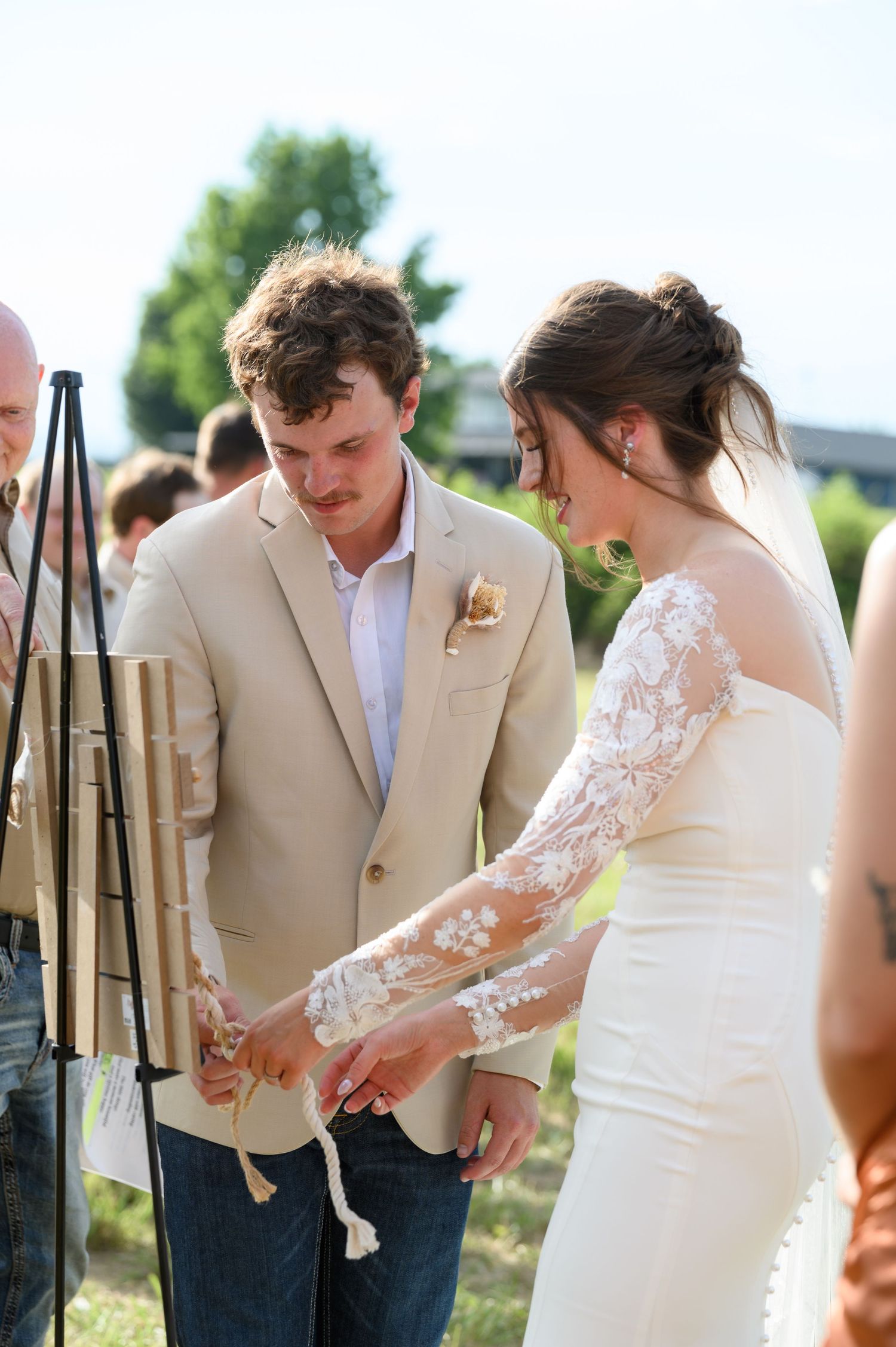 Bride and groom in tan suit participate in a cord of three strands unity ceremony outdoors.