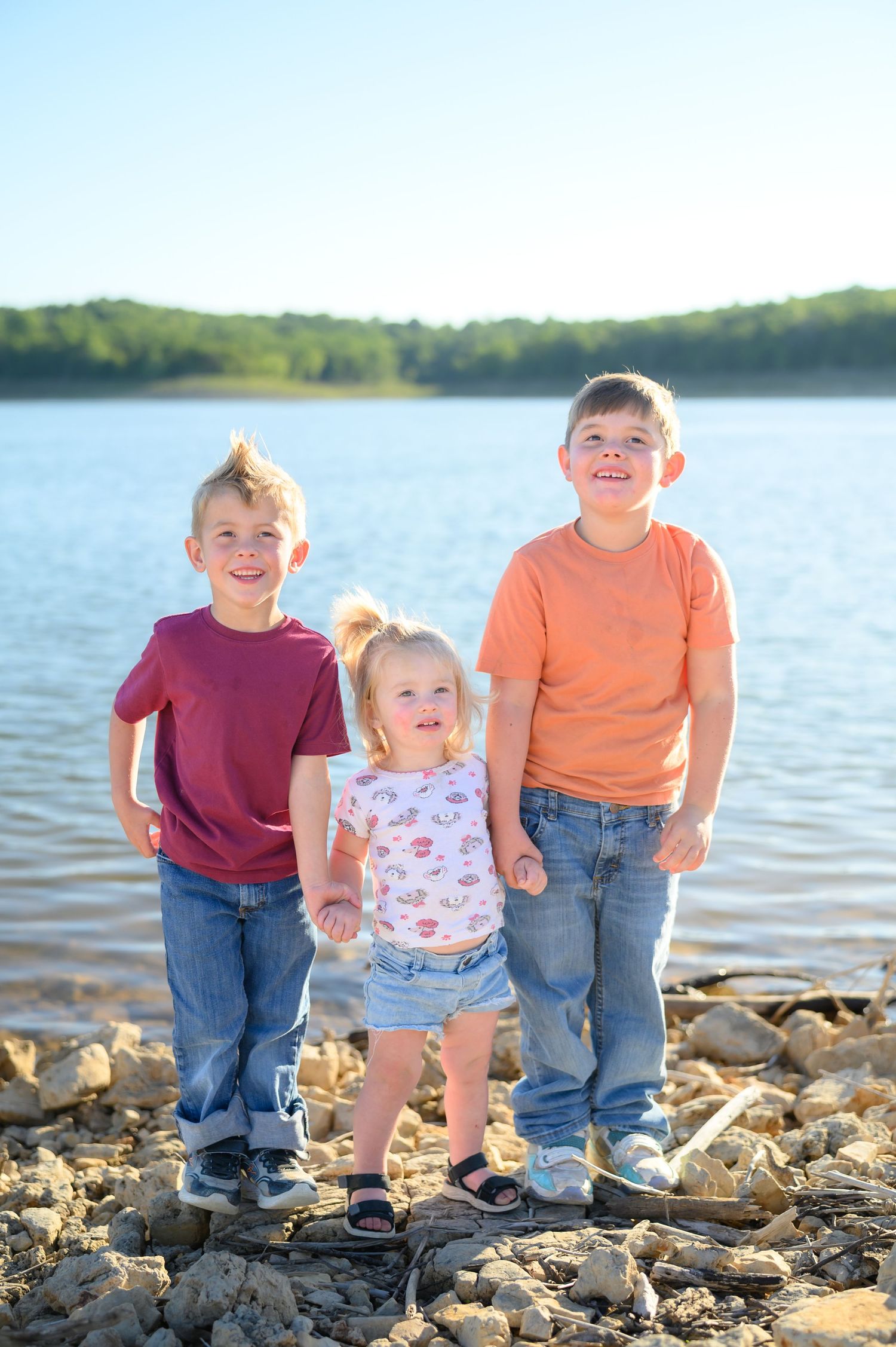 Three young siblings standing on rocky lakeshore smiling, with lake and green trees in background.