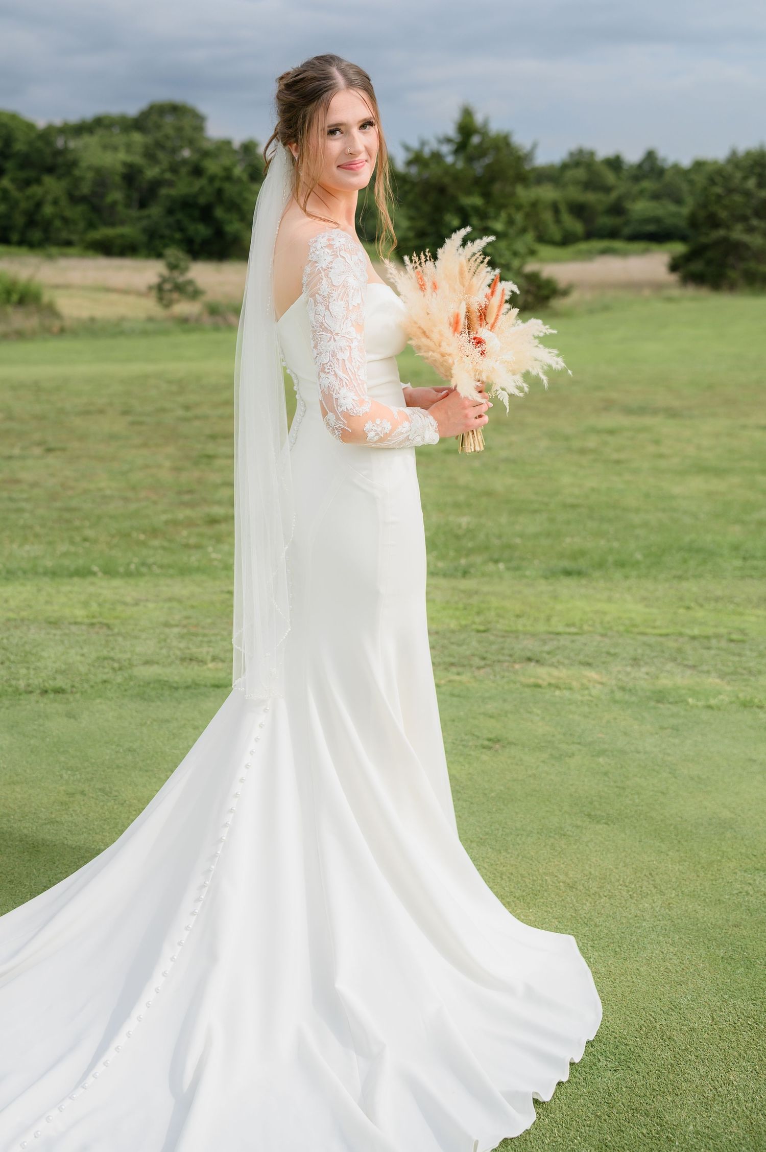 Elegant bride in long-sleeve lace wedding gown with cathedral veil holding a pampas grass bouquet outdoors.