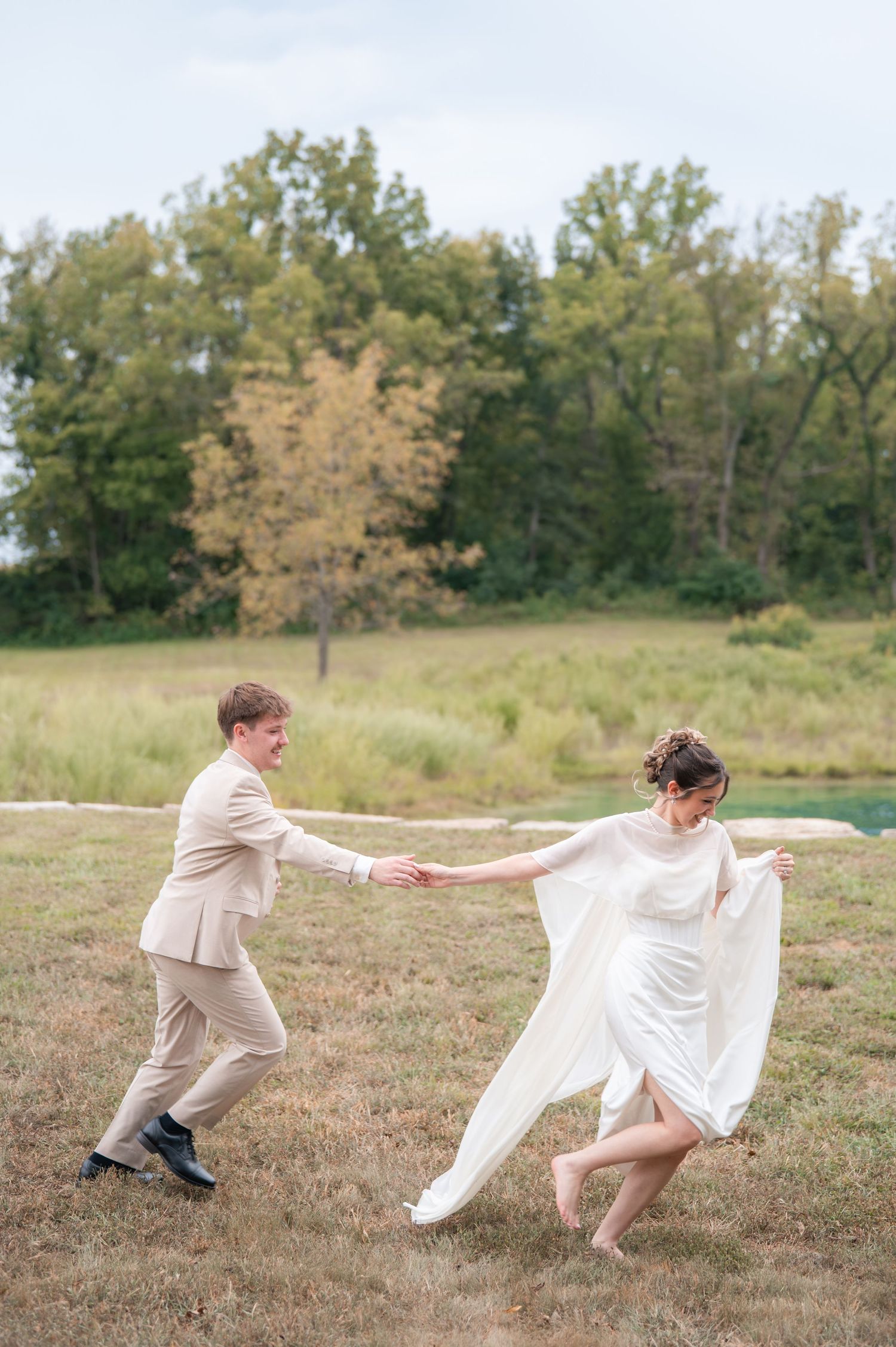A bride and groom dance joyfully in a field, her white gown flowing, with lush green trees in the background.