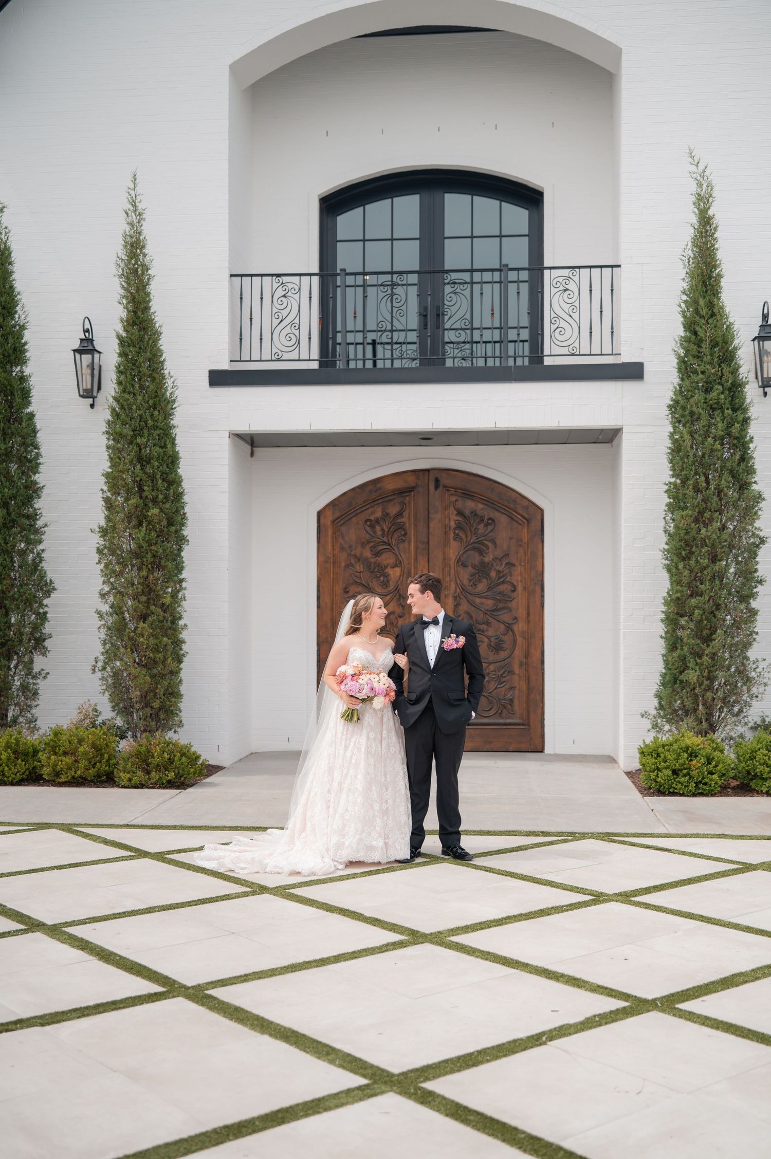 Bride and groom posing together outside elegant white wedding venue with wood doors.