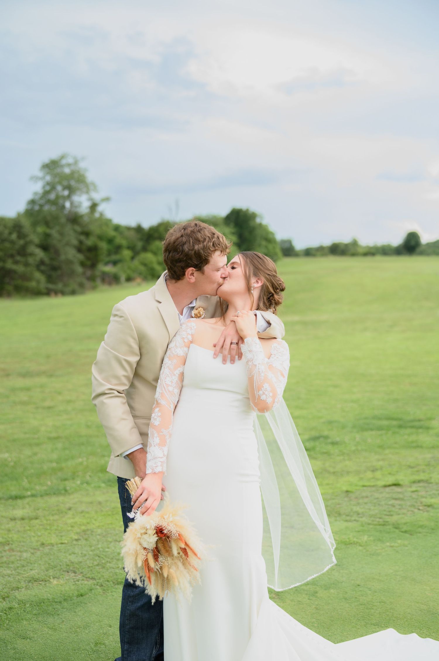 Bride and groom sharing a kiss on a green lawn, groom holding pampas grass bridal bouquet.