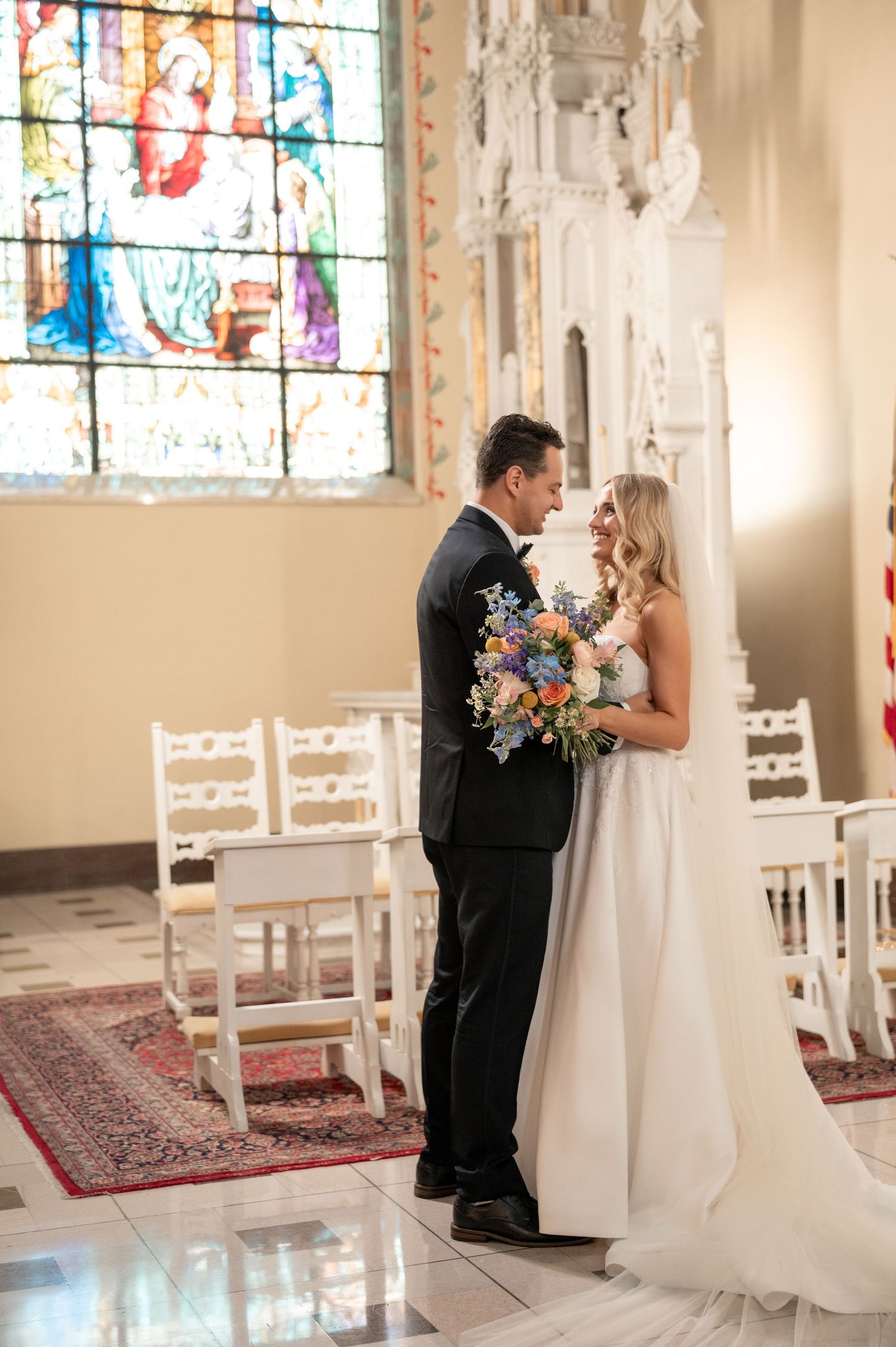 Bride and groom share an intimate moment inside a stunning white church with vibrant stained glass windows.