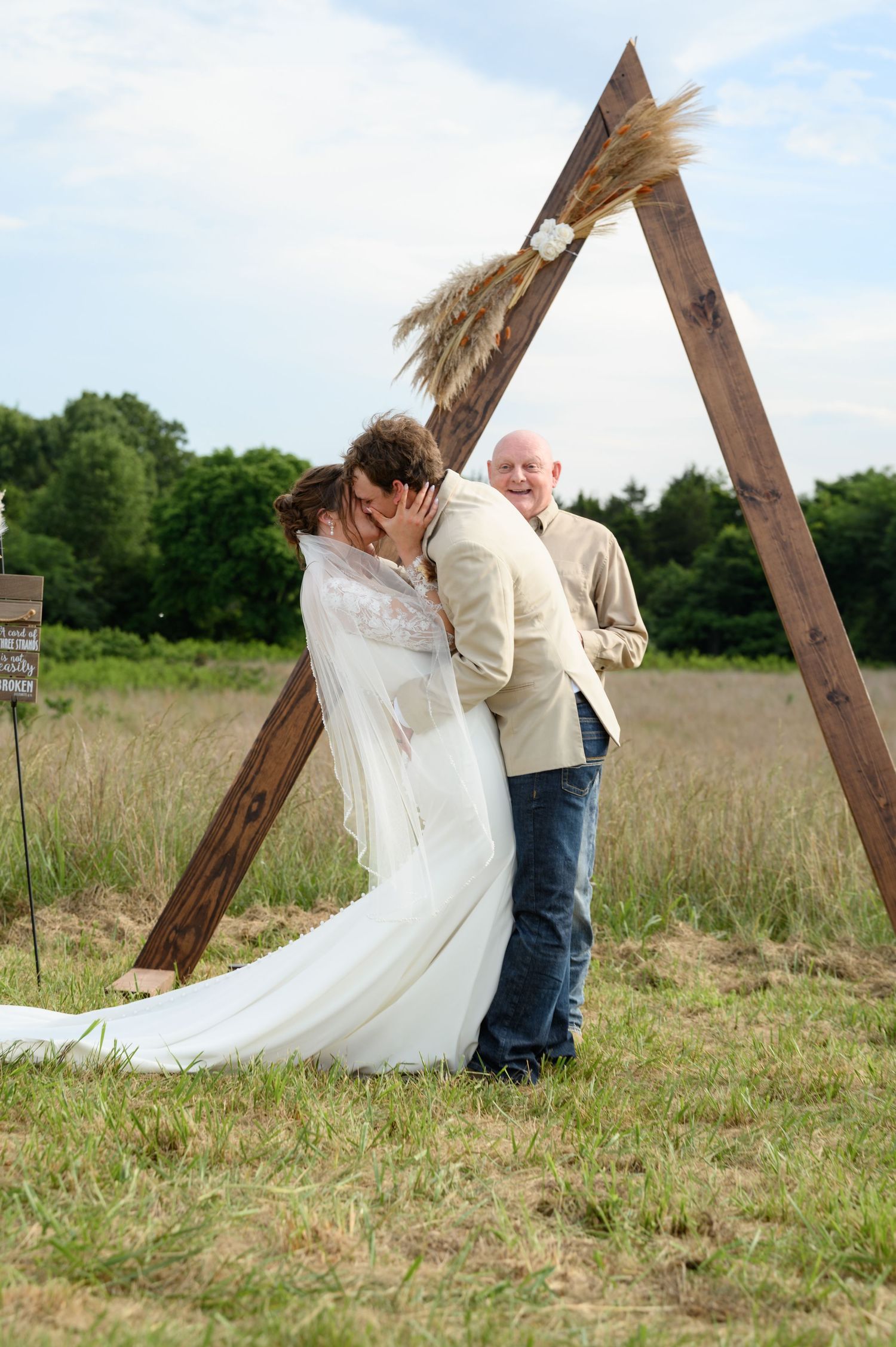 Newlyweds share first kiss under a wooden triangular arch decorated with pampas grass at outdoor wedding.