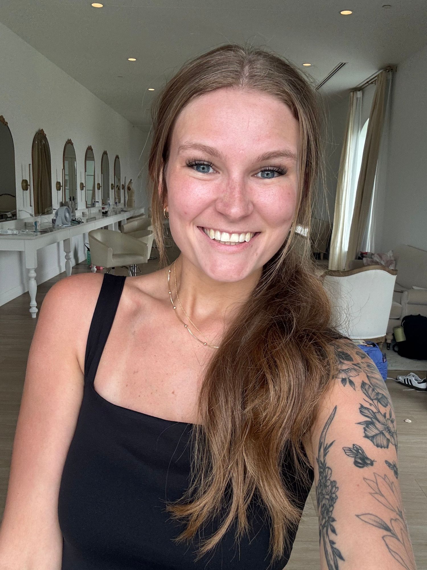 Smiling woman with long brown hair and floral arm tattoo wearing black top in bright salon setting.