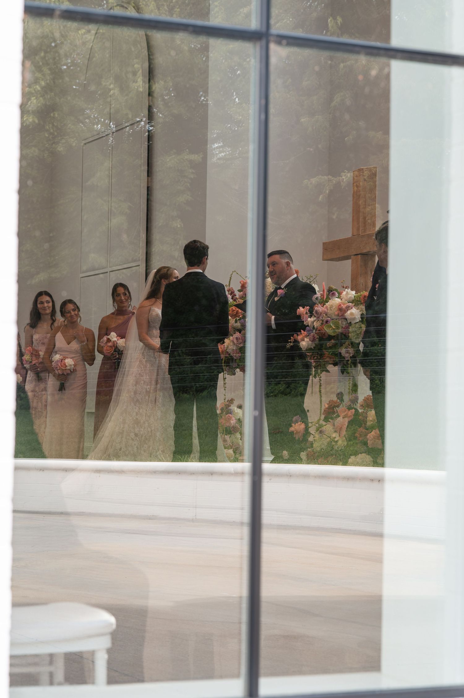 Wedding ceremony reflected in chapel window with bridesmaids and floral arrangements.