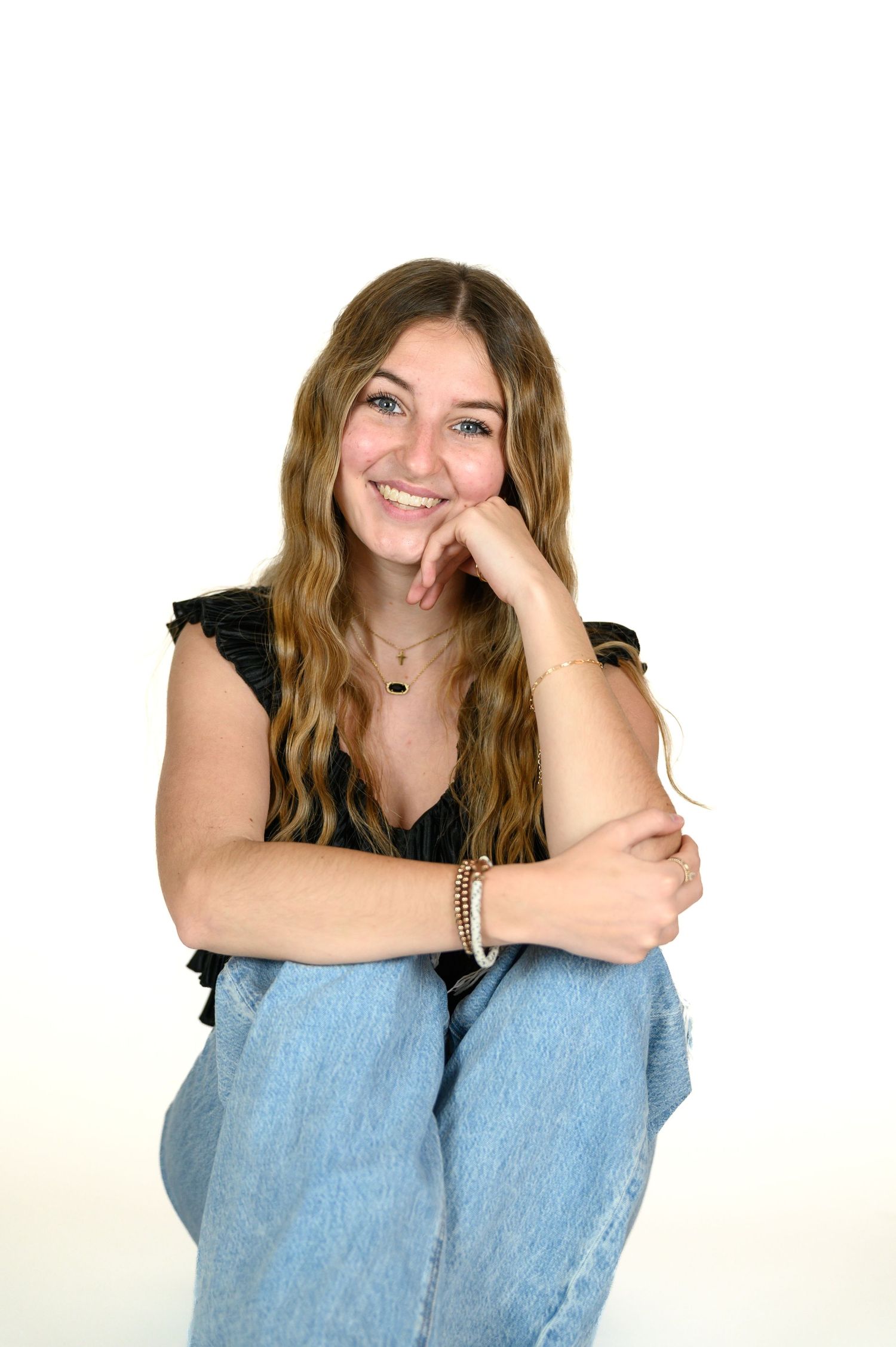 Smiling teenage girl with long blonde hair sitting casually in jeans and black top against white background.
