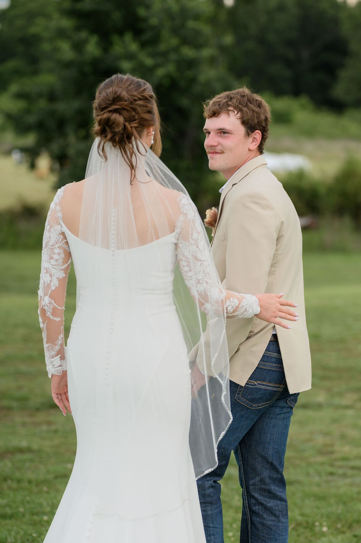 Groom in tan blazer smiles at bride in white lace wedding gown during outdoor first look.