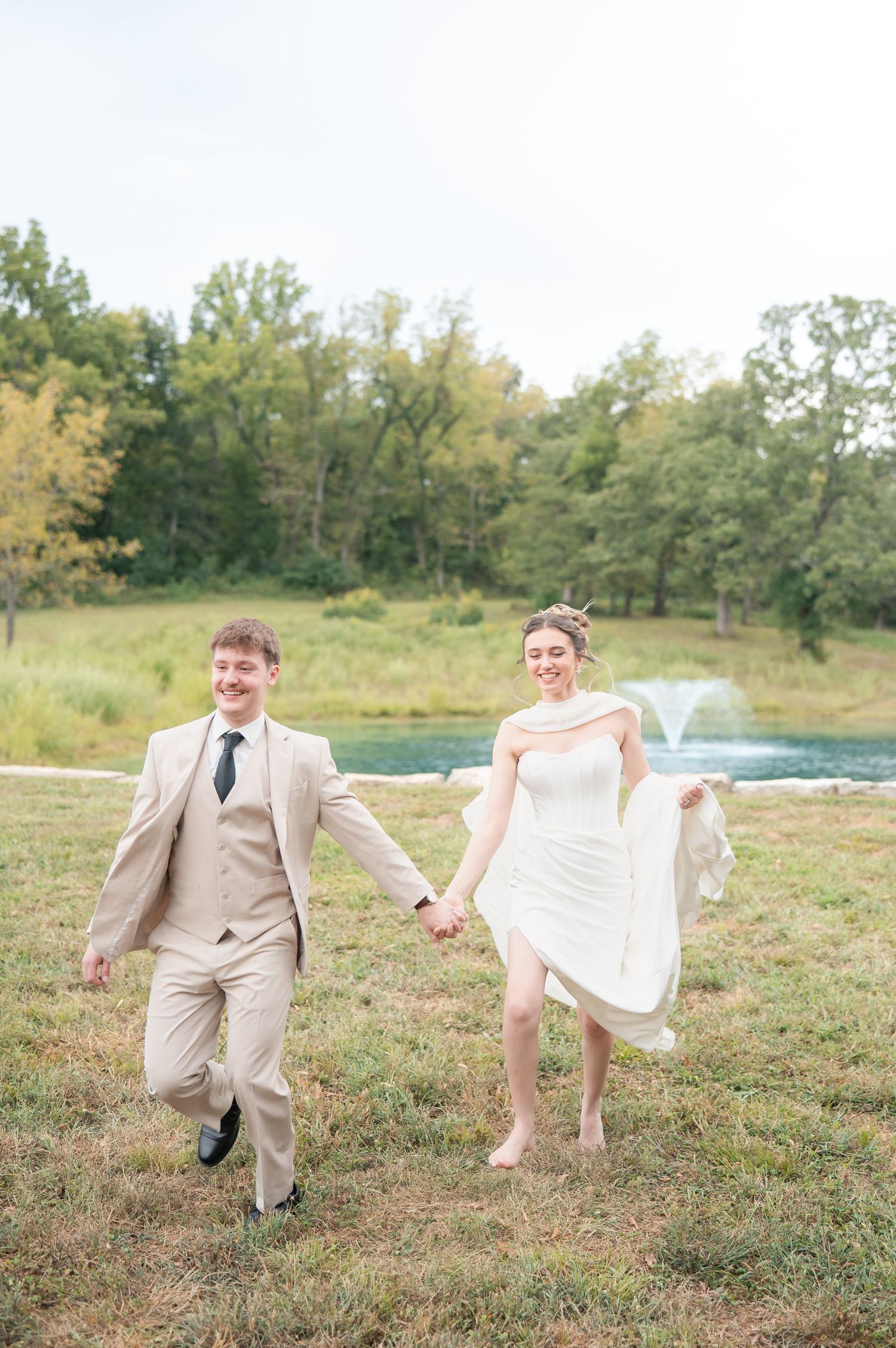 A joyful bride and groom hold hands while running through a lush green lawn near a pond on their wedding day.