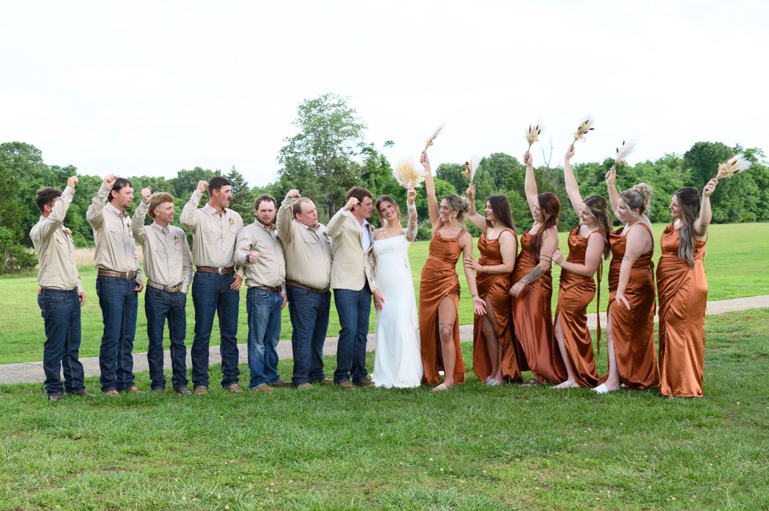 Wedding party with bridesmaids in burnt orange satin gowns and groomsmen in tan suits cheering outdoors.