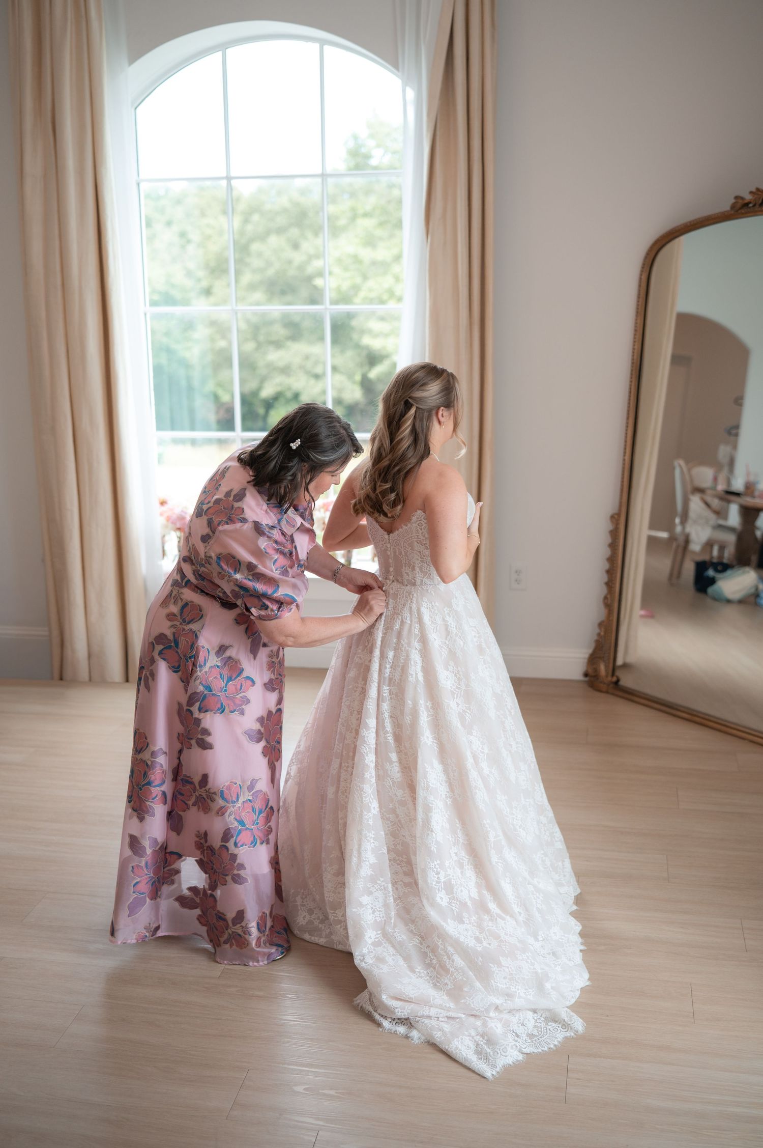 Mother helping bride fasten lace ballgown wedding dress in bright bridal suite with gold mirror.