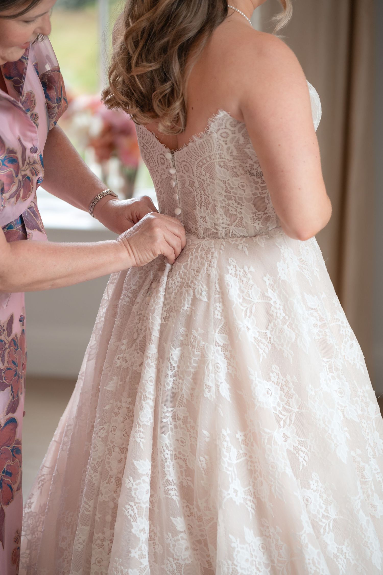 Mother buttoning back of brides blush lace wedding gown before ceremony.