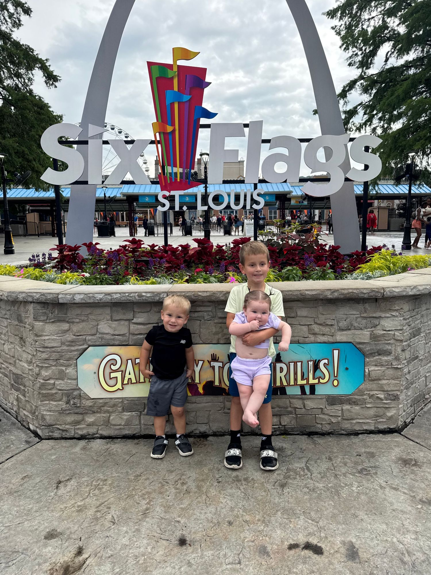 Three children pose at Six Flags St. Louis entrance sign with Gateway Arch replica and colorful park decor.