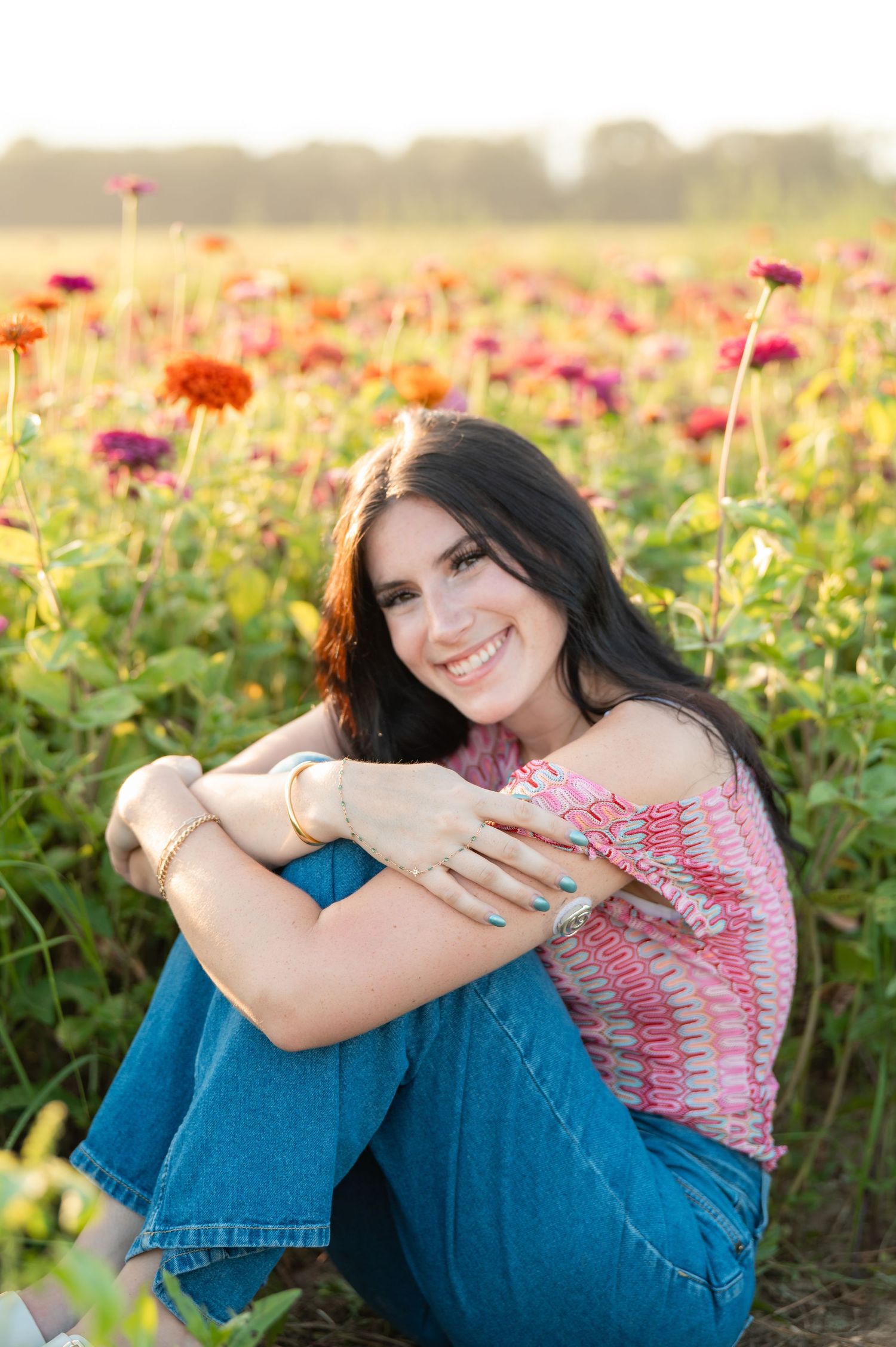 Young woman smiling in a colorful zinnia flower field at golden hour, wearing pink top and jeans.